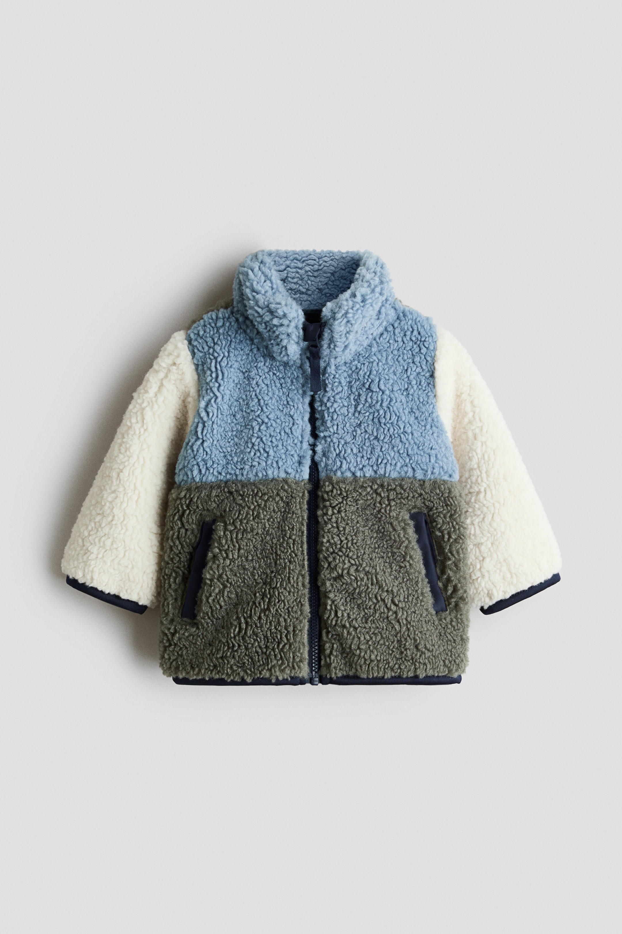View larger image: A colour-blocked teddy jacket is shown from the front, featuring light blue on the upper front and stand collar, olive green on the lower front, and cream sleeves. A navy zipper runs down the front, with diagonal front pockets and navy jersey bindings finishing the cuffs and hem.
