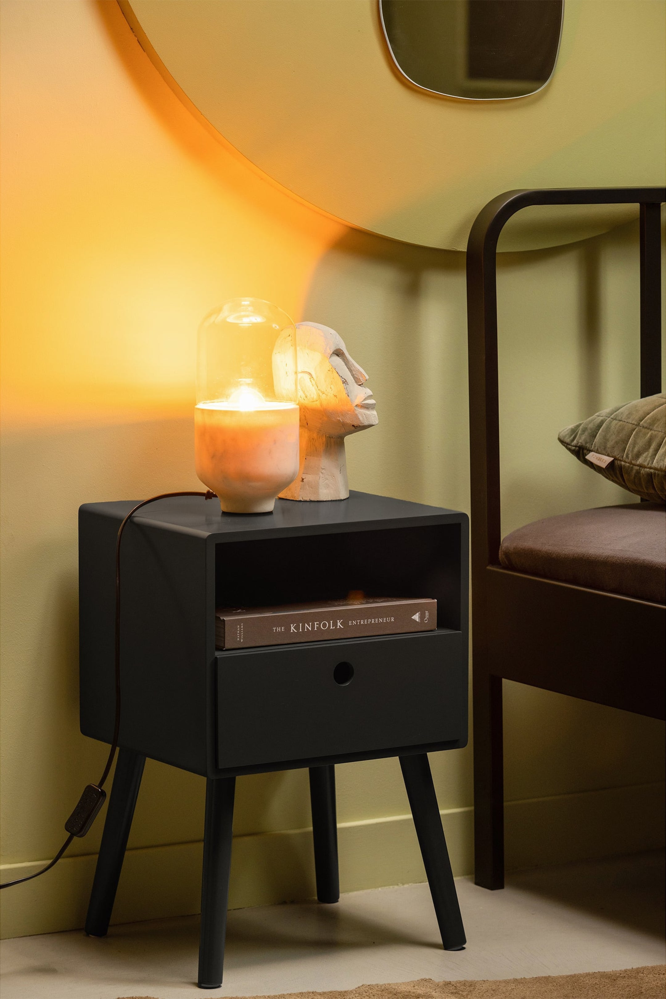 Grotere afbeelding bekijken: Een zwart nachtkastje toont zijn voor- en linkerkant, met een open plank en een lade met een ronde uittrekgreep. Het heeft een boek op de plank en een lamp en een hoofdsculptuur op het bovenoppervlak, staande op vier taps toelopende poten.