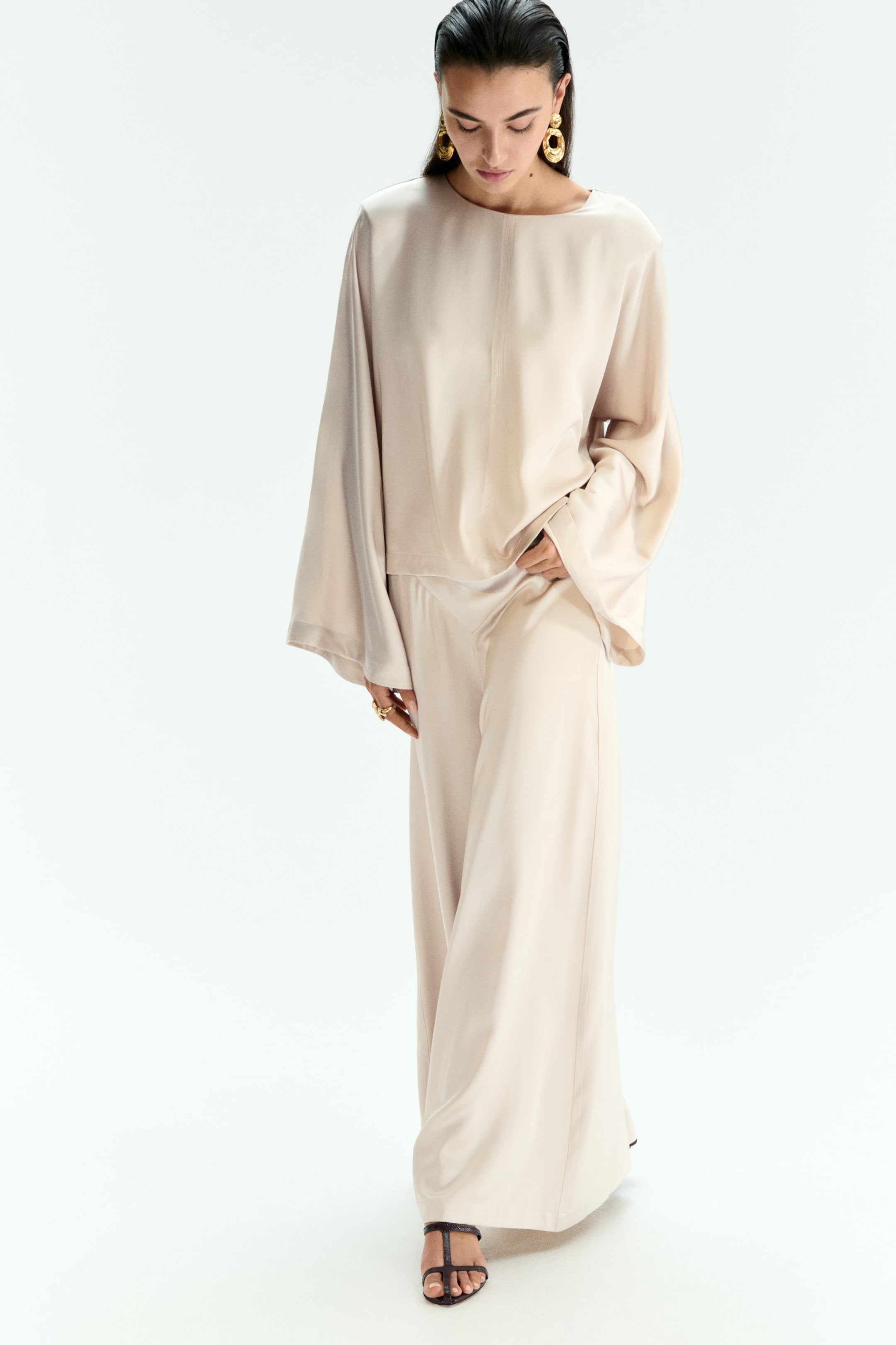 View larger image: A light beige matching set, consisting of a boat-neck top with wide, flowing sleeves and a cropped hem, and wide-leg trousers, is being worn by the model, who is resting one hand at their hip, with fingers touching the top's hem and the trousers' waistband.