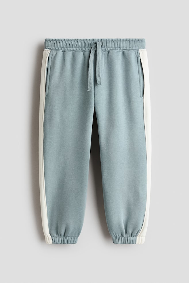 A pair of light blue-grey sweatpants is shown from the front, featuring an elasticated ribbed waistband with an adjustable drawstring and side pockets. Off-white contrast stripes are running down the sides of each leg, which is tapering into elasticated cuffs. - 1