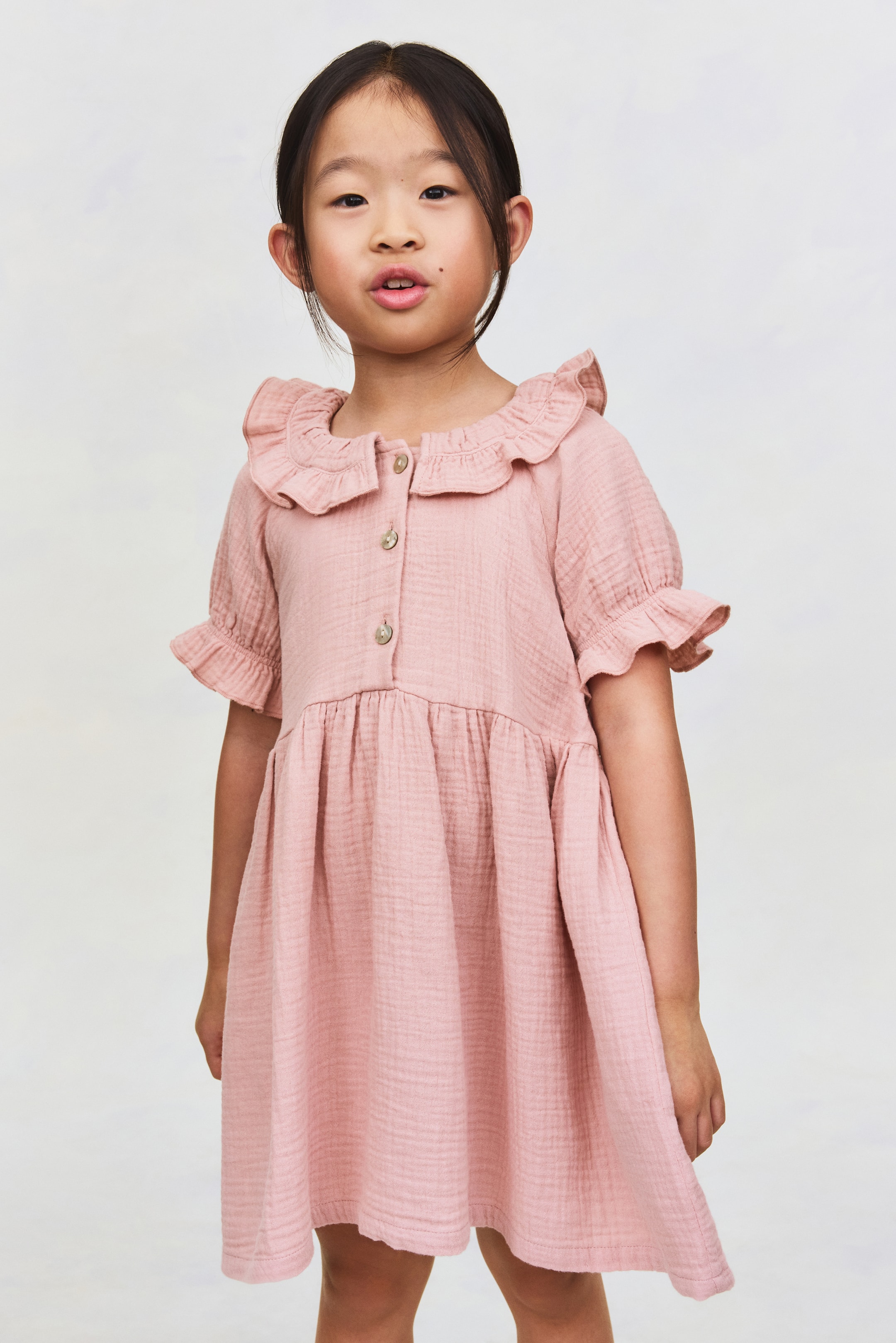 View larger image: A pink crinkled dress features a ruffled collar, a three-button placket and short puffed sleeves with ruffled cuffs, with a gathered seam creating a flared skirt.