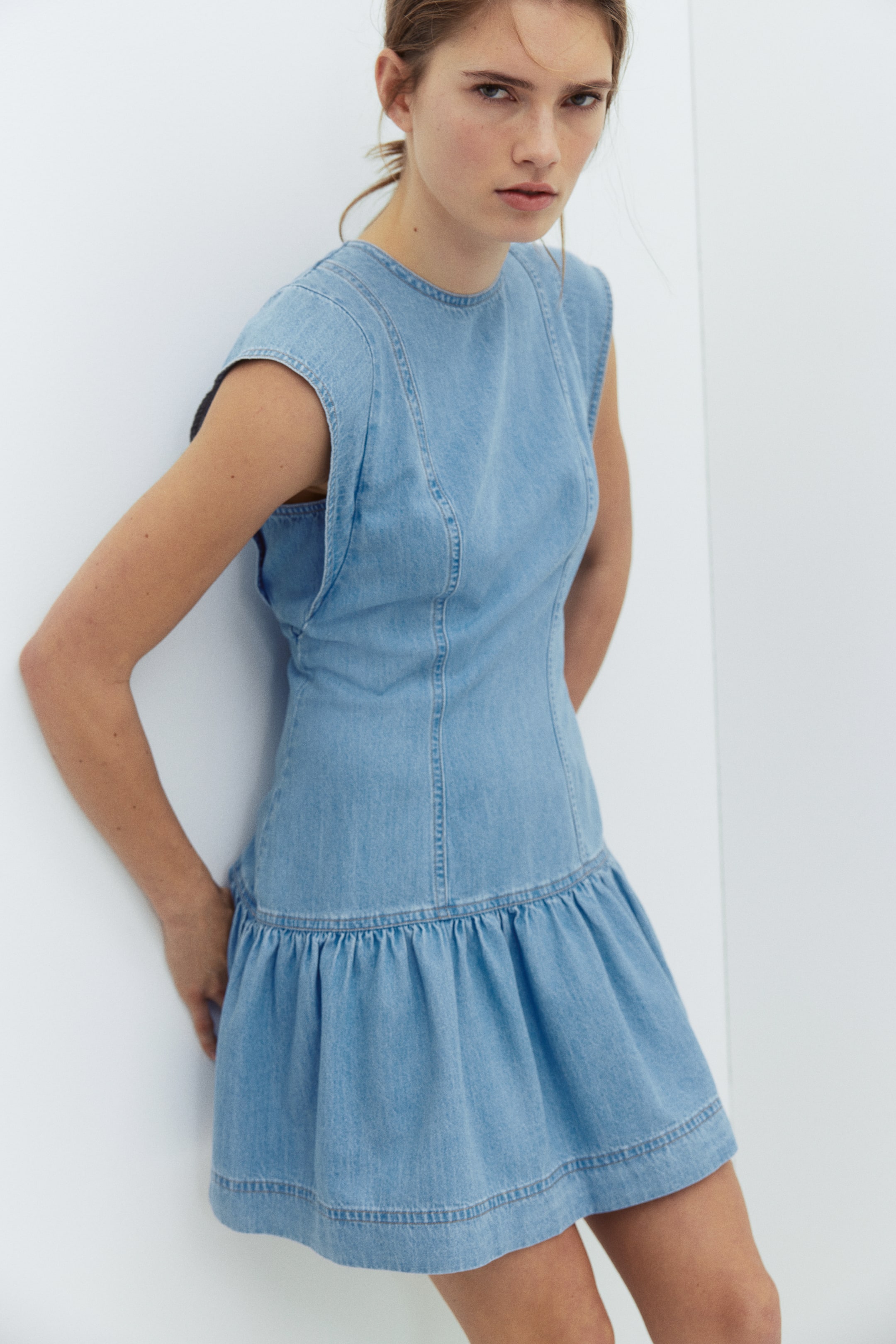 查看大圖: 淺藍色牛仔連身裙展示修身上身，設有縱向車線、圓領及無袖剪裁；腰部收褶，形成喇叭形裙襬，下襬以雙線車縫處理。畫面以前左方角度呈現。