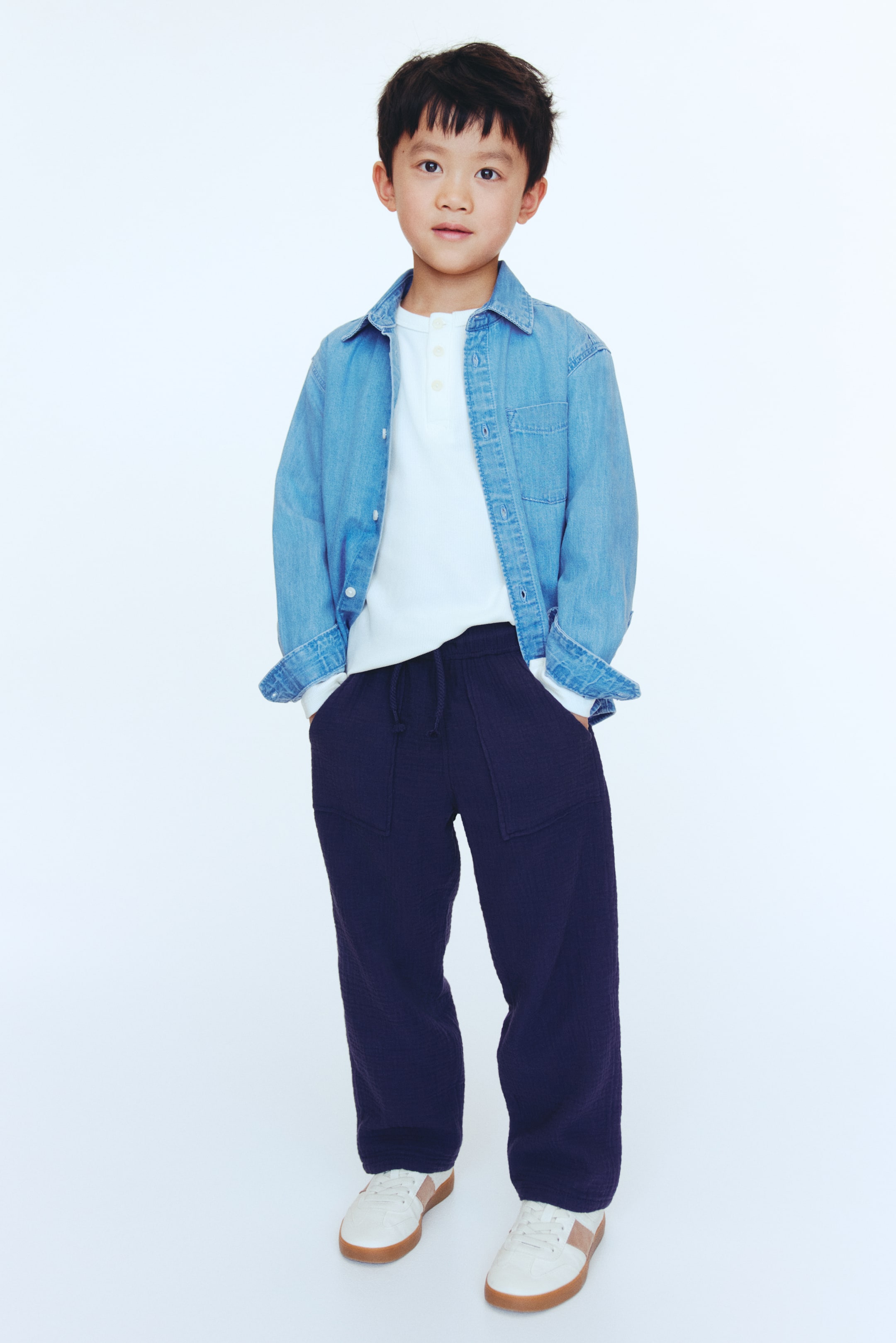 View larger image: A child's outfit features a light blue denim shirt, unbuttoned with rolled-up sleeves, layered over a white long-sleeve Henley shirt with a three-button placket. Dark blue textured pants with a drawstring waist and large front patch pockets complete the look, along with white sneakers featuring light brown accents and gum soles.