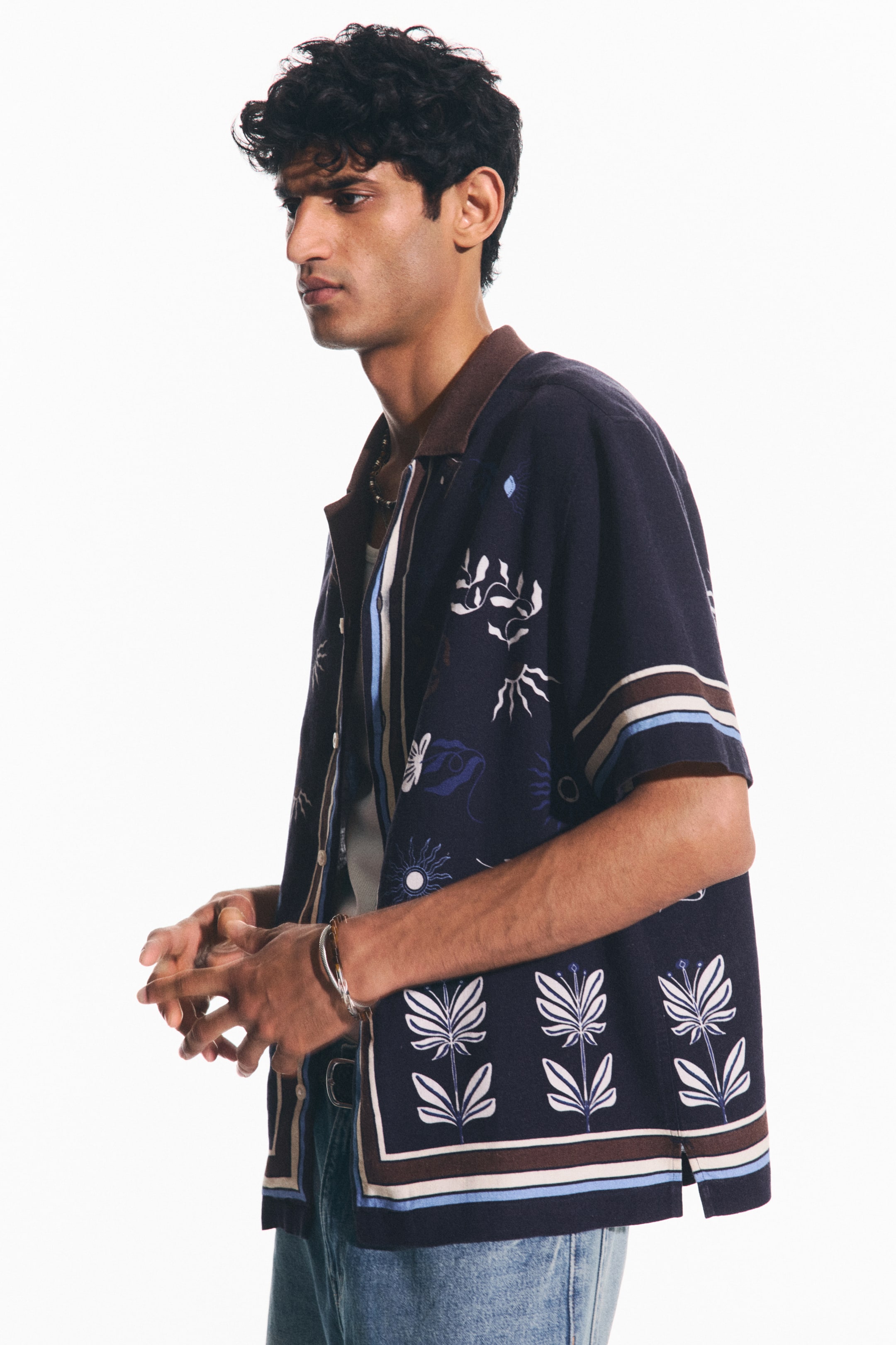 View larger image: The model is sporting a short-sleeved, navy printed shirt adorned with a brown resort collar, showcasing white and light blue botanical patterns. It is styled open over a light undershirt. The shirt is designed with horizontal stripes on the sleeves and features a straight-cut hem. The model is posing with clasped hands.