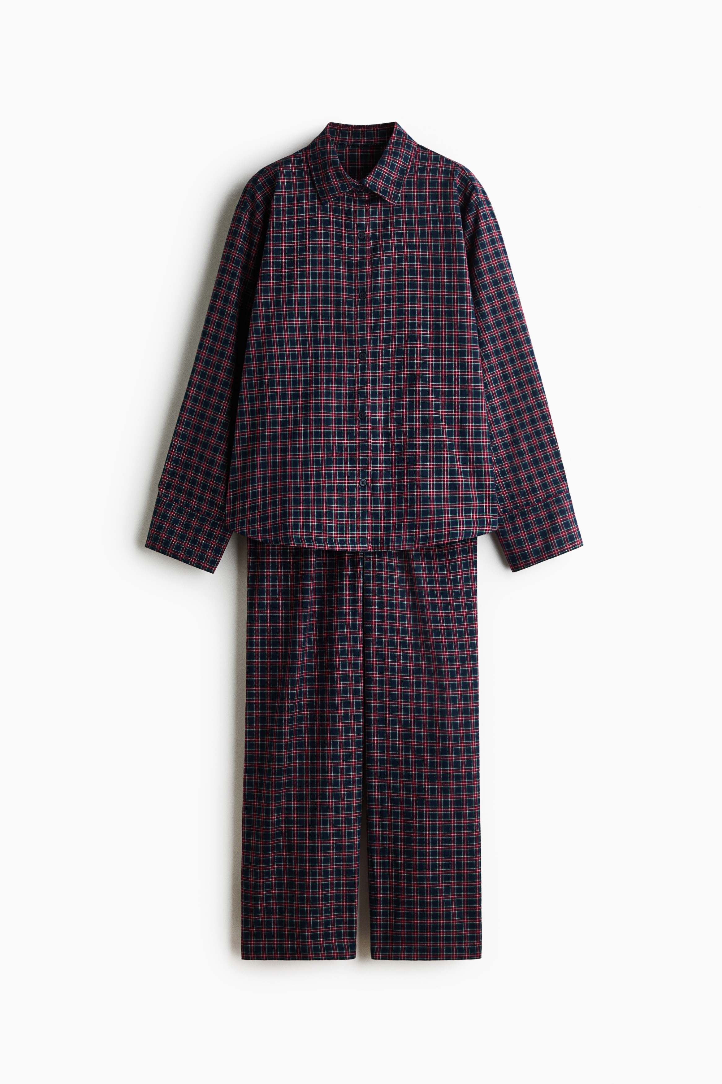 A red and dark blue checked cotton flannel pyjama set is being worn by the model seated cross-legged. The long-sleeved shirt features a collar and a button-front fastening, worn partially open with the sleeves pushed up, while the matching wide-leg trousers complete the set.