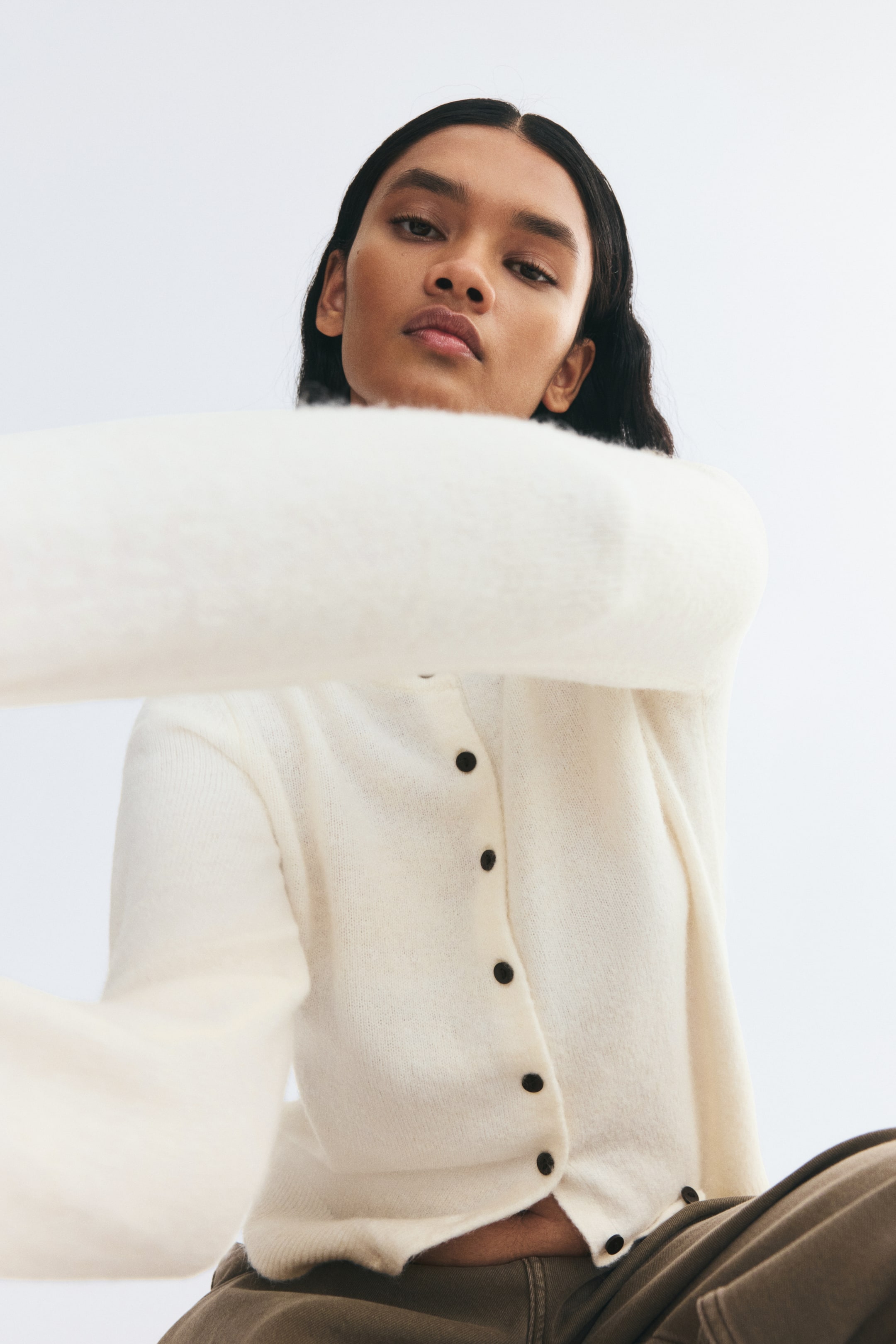 View larger image: A cream-colored soft-knit cardigan is worn by the model, who is raising one long sleeve toward the foreground. The fitted cardigan features dark buttons down the front and a ribbed hem, and is styled with casual brown trousers.