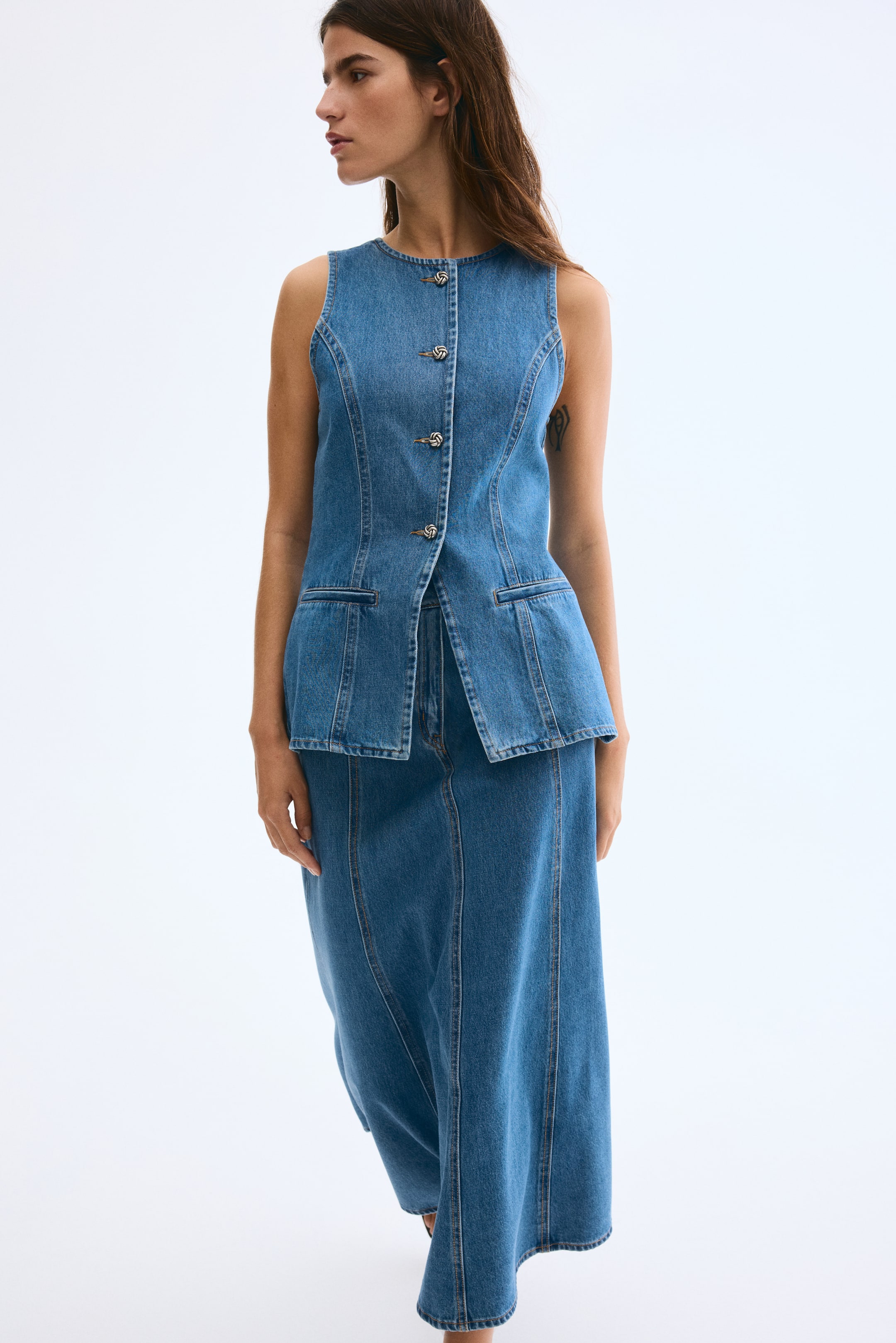 View larger image: A sleeveless denim vest is featuring a round neckline, three decorative twisted silver-toned buttons down the front, and two flap pockets at the hips. It is paired with a long denim skirt that showcases vertical seam detailing and a flared silhouette.