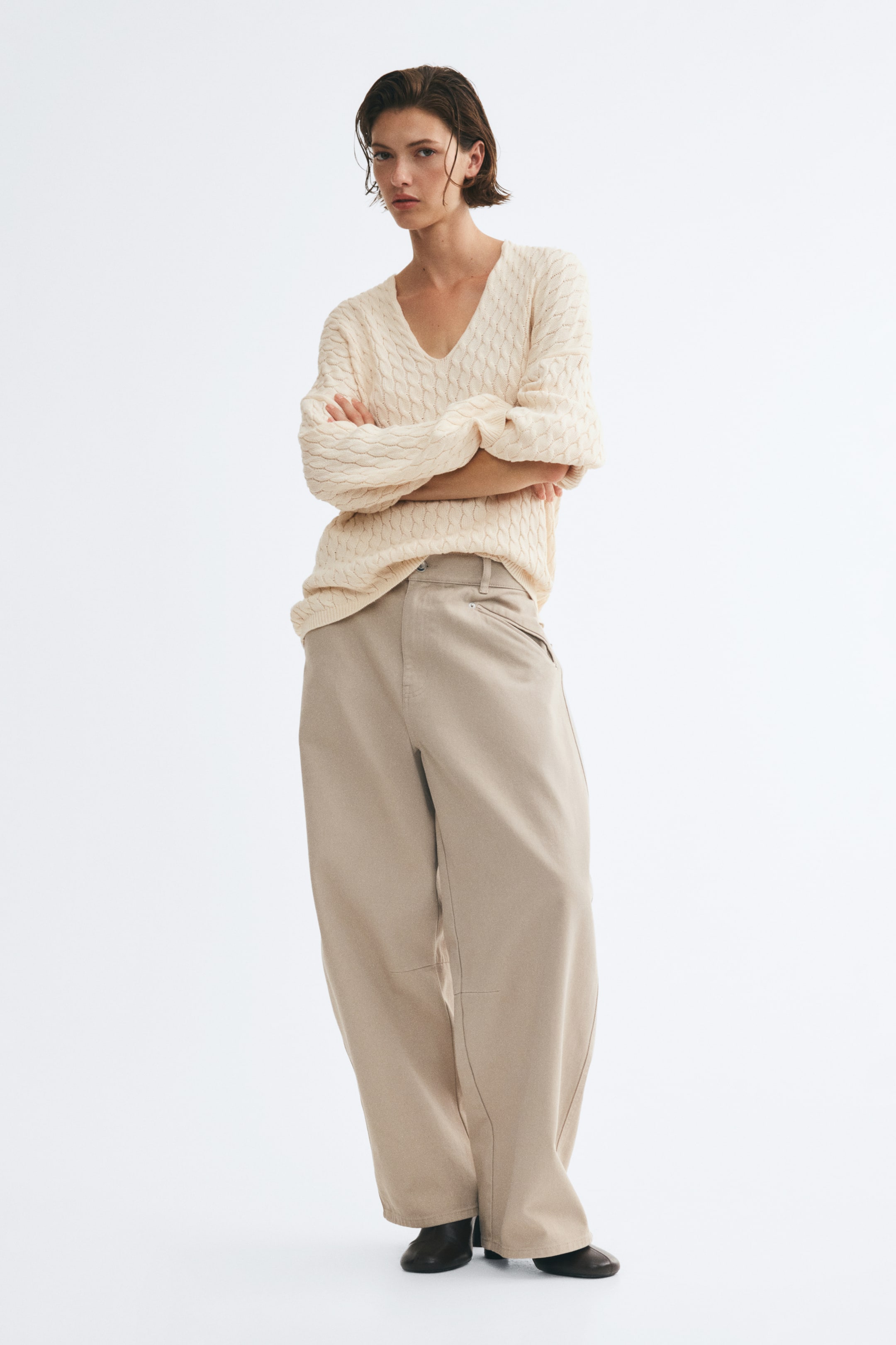Visualizza immagine più grande: Un maglione oversize color crema, lavorato a trecce, con profondo scollo a V e spalle scese, indossato dalla modella. La modella è in piedi con le braccia incrociate, abbinando il maglione a pantaloni beige a gamba larga.