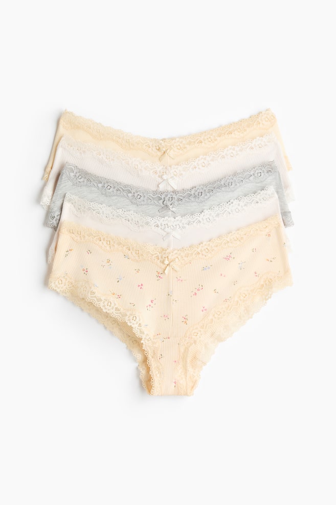Four ribbed cotton jersey briefs are stacked, presented front-facing, each featuring lace trims along the low, V-shaped waist and leg openings, and a small bow at the center front. The set includes light yellow, off-white, light grey, and a light yellow brief with a floral print. - 1