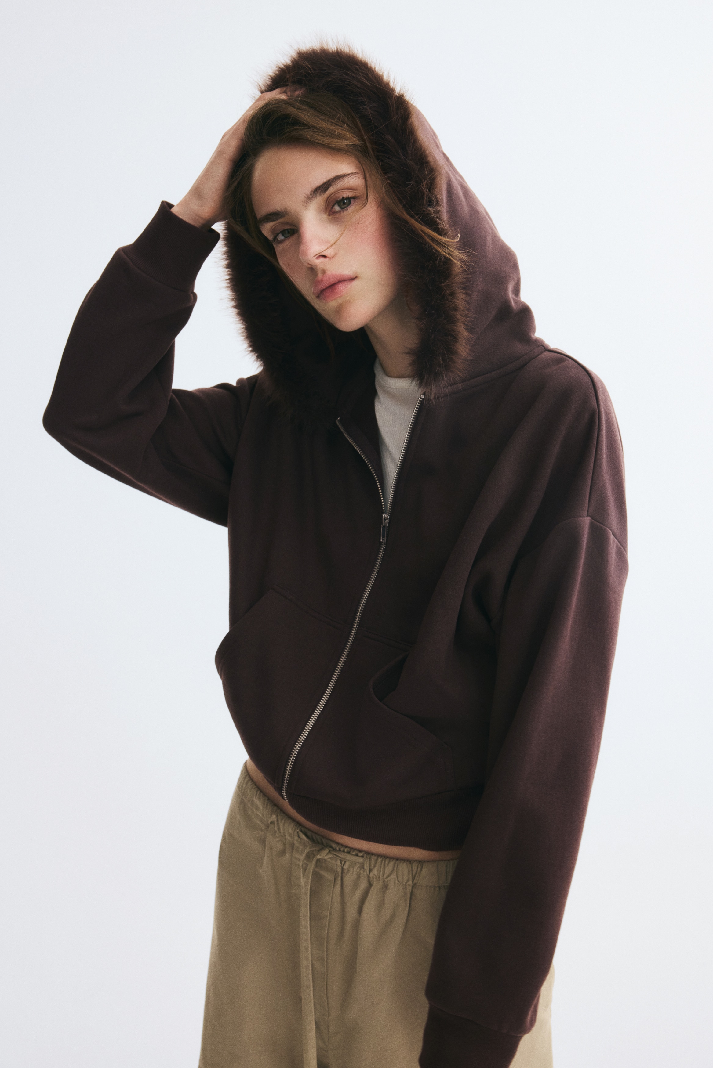 A dark brown zip-up hoodie is shown from the front, featuring a plush faux-fur trim lining the hood. The design features a silver-toned metal zipper, a split kangaroo pocket, dropped shoulders, and ribbed cuffs and hem.