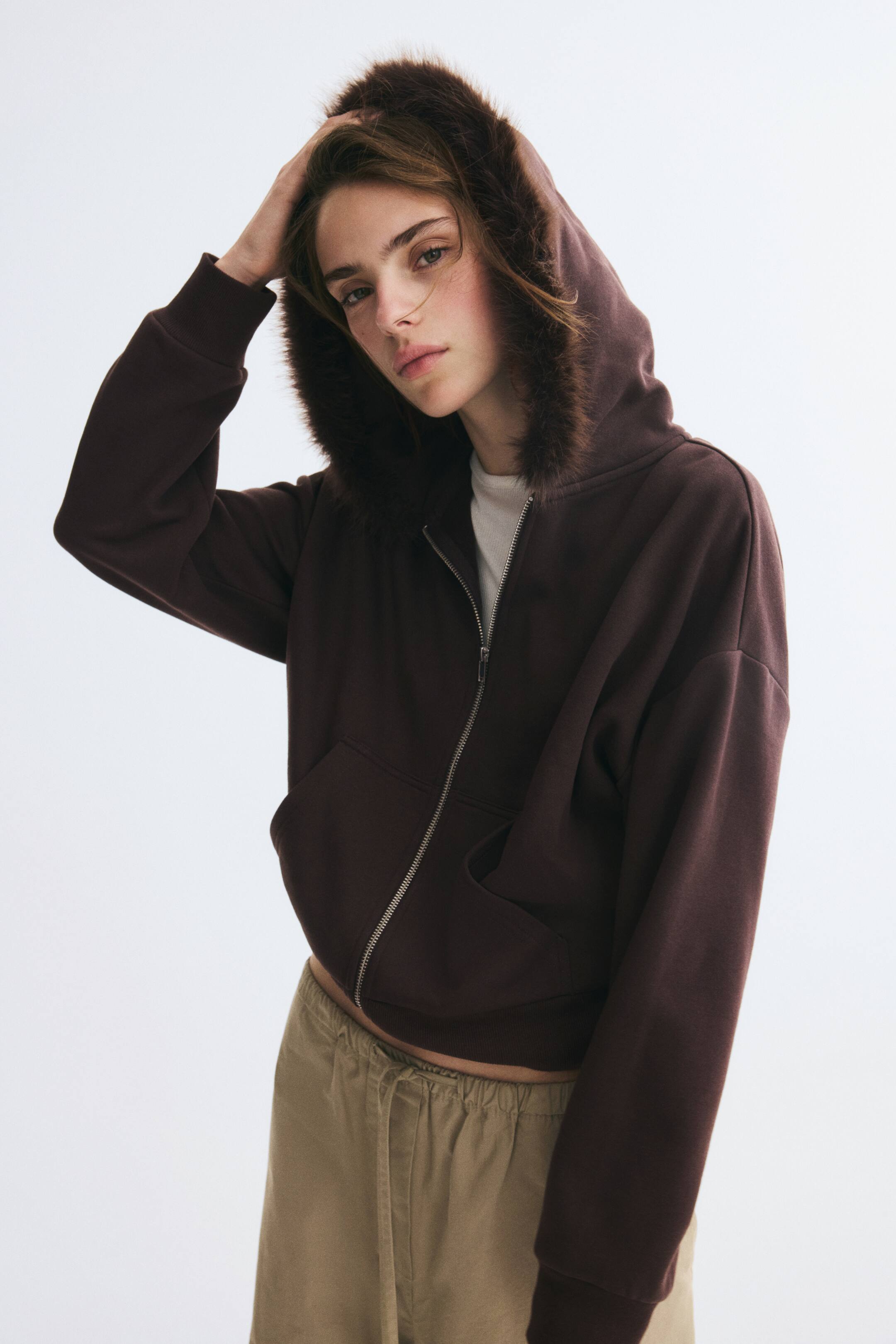 View larger image: A dark brown zip-up fleece hoodie featuring a hood with a fluffy faux-fur trim is being worn by the model, who is adjusting the top of the hood with one hand. The loose-fit hoodie has dropped shoulders, front pockets, and ribbed cuffs, and is styled over a white T-shirt and beige drawstring trousers.