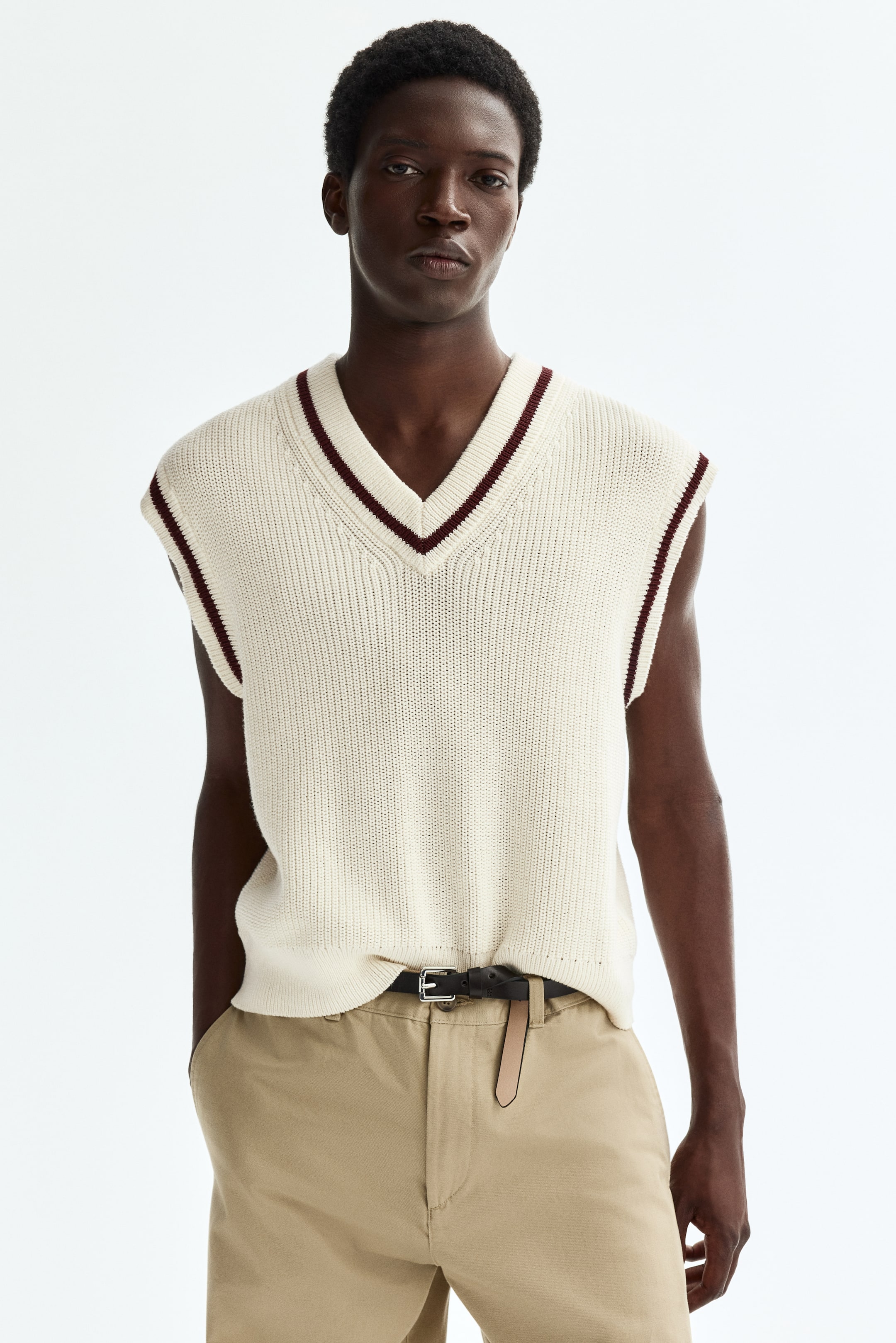 View larger image: A cream sweater vest features a ribbed knit texture and a V-neckline with burgundy and cream striped trim along the neck and armholes. It fits loosely on the torso and is slightly tucked into the waistline at the front.