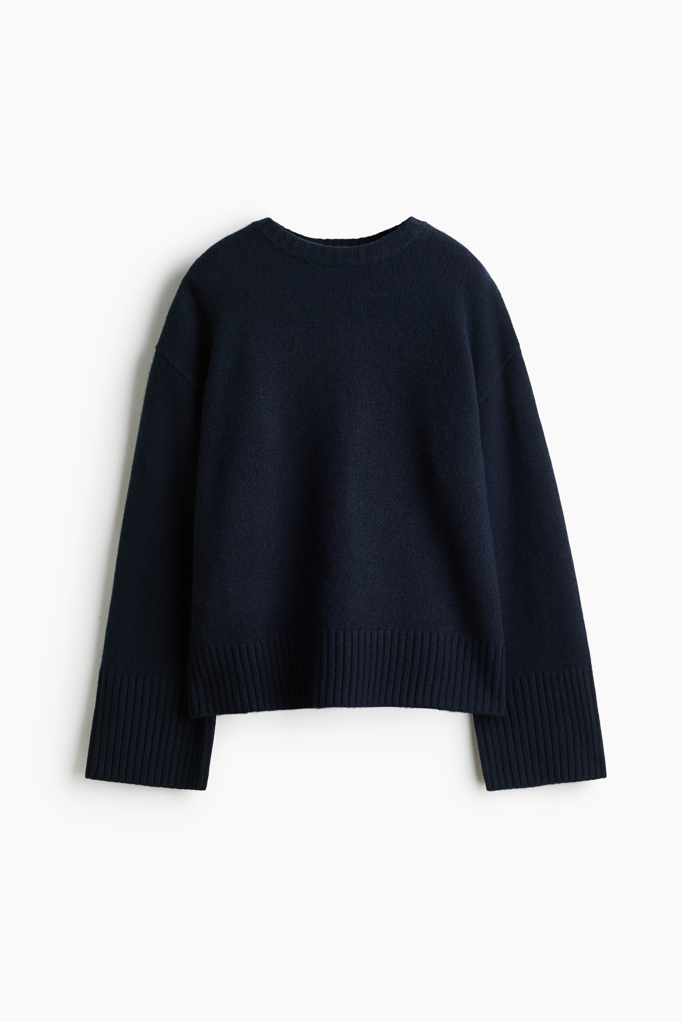 En oversized mÃ¶rk marinblÃ¥ stickad trÃ¶ja bÃ¤rs av modellen tillsammans med blÃ¥ jeans. TrÃ¶jan har nedhasade axlar och extra lÃ¥nga Ã¤rmar med breda ribbade muddar och en ribbad fÃ¥ll.