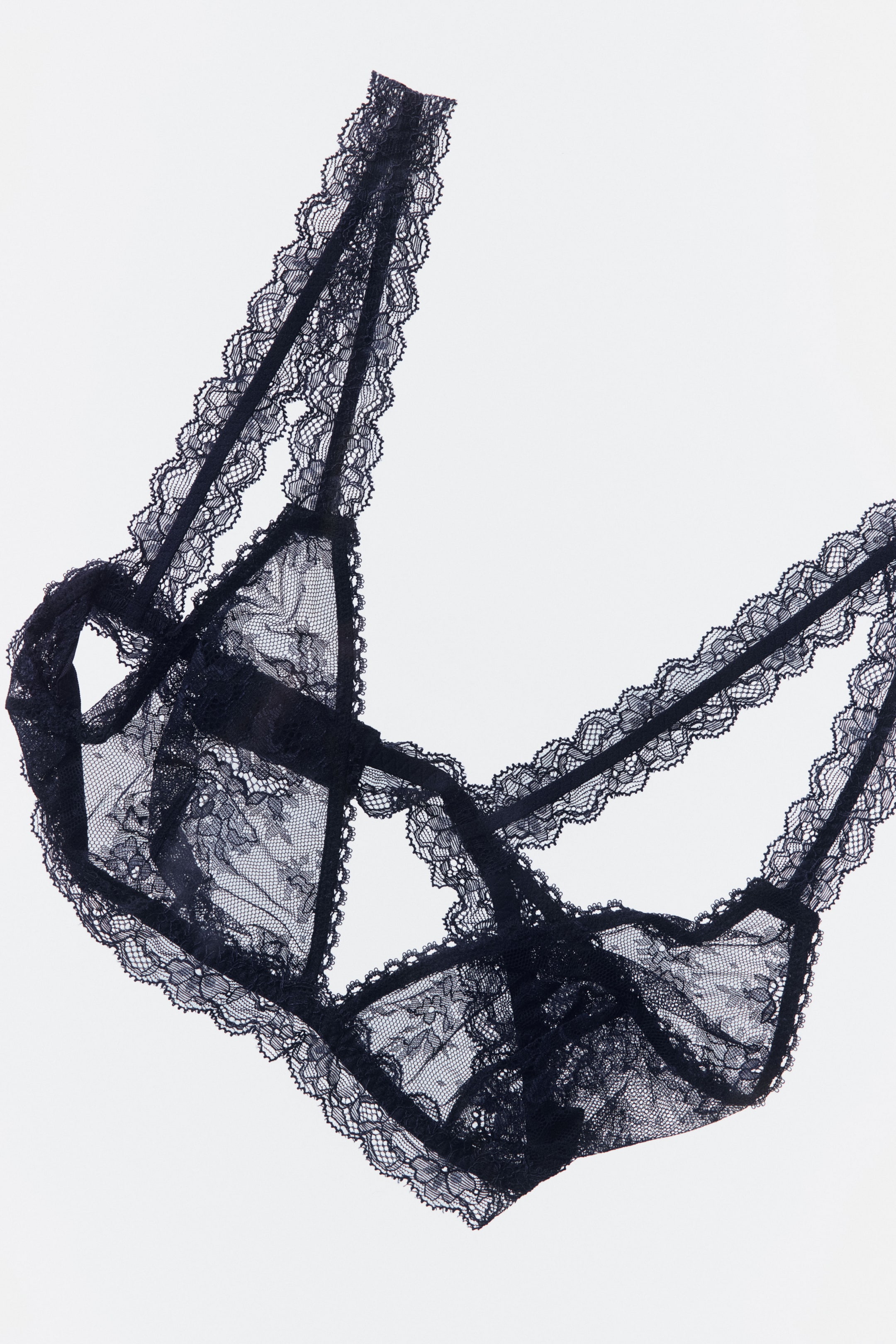 Größeres Bild ansehen: Ein schwarzes Spitzen-Bralette liegt flach da und präsentiert seine transparenten, floralen Spitzen-Dreieckskörbchen. Mehrere zarte Träger verbinden die Körbchen und bilden ein käfigartiges Design, während eine gewellte Spitzenborte alle Kanten umrahmt.