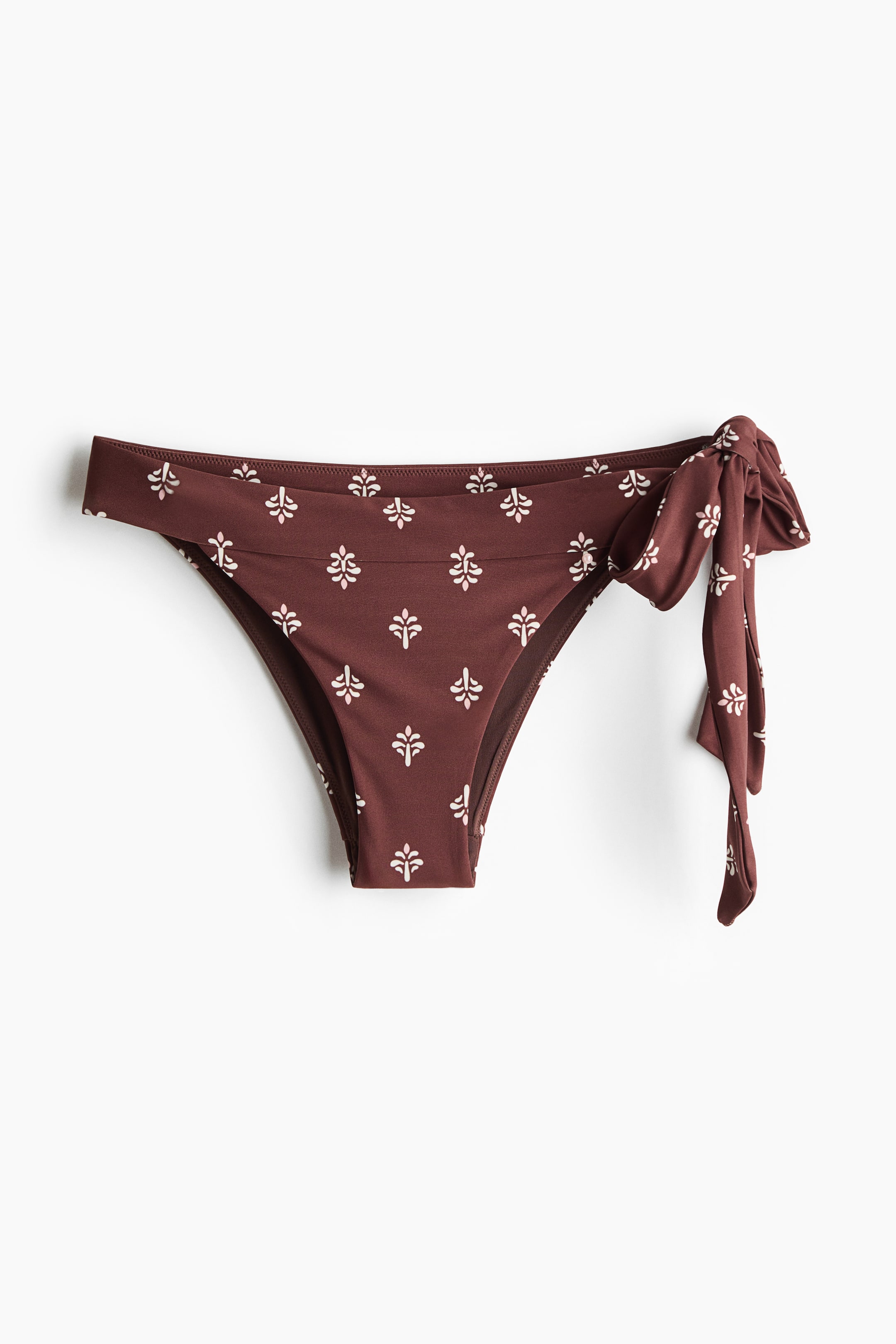 View larger image: The fully lined bikini bottoms are shown from the front, featuring a dark brown base with an all-over off-white floral pattern. They are crafted with a mid-waist design, showcasing wide ties knotted at the right side, and provide cutaway coverage at the back.
