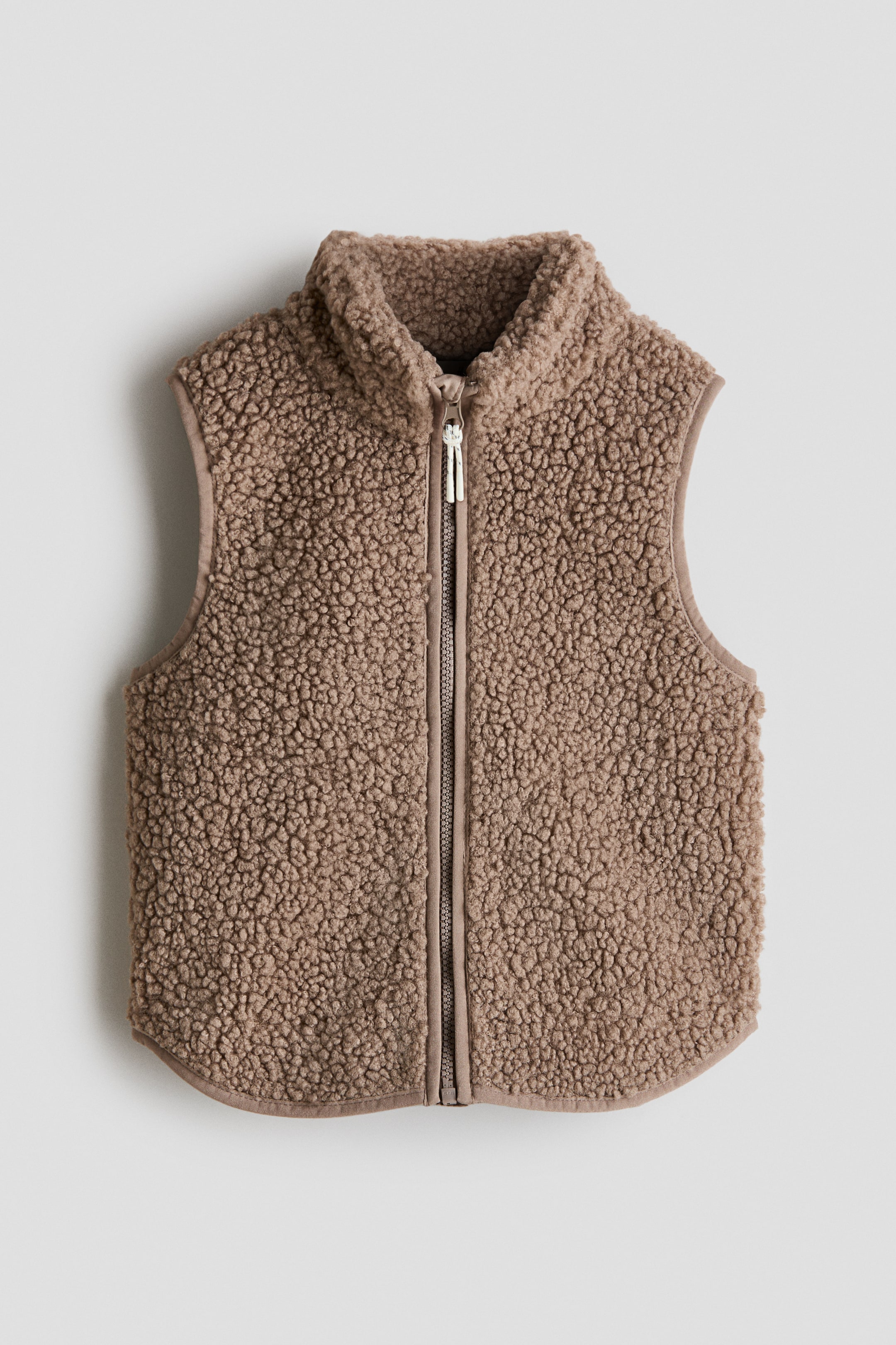Grotere afbeelding bekijken: Een bruine teddy fleece bodywarmer is frontaal gericht en heeft een ritssluiting met een zilverkleurige ritstrekker, een opstaande kraag en een licht gebogen zoom met bijpassende afwerking rond de armsgaten en onderkant.