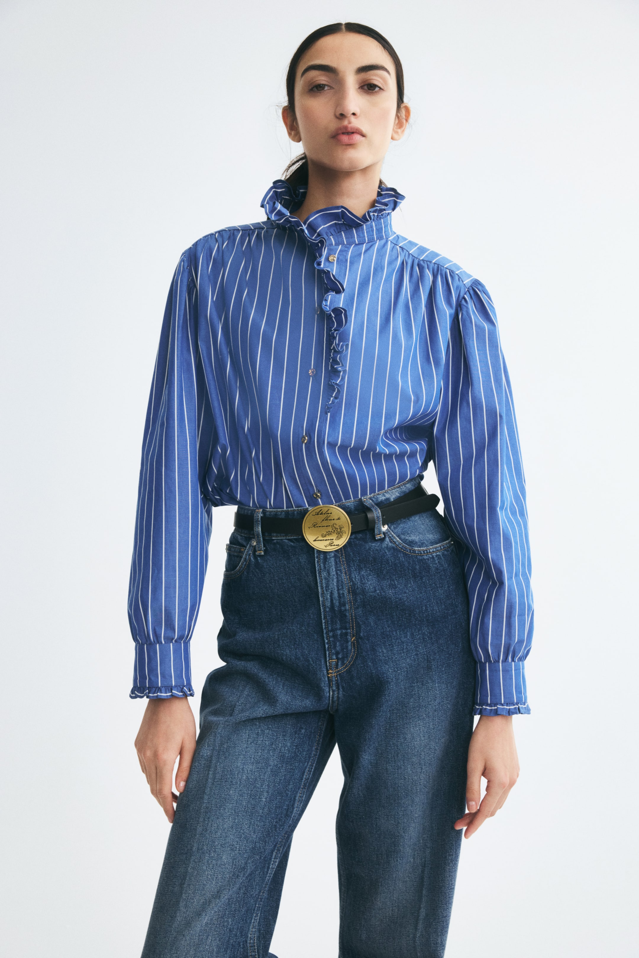 View larger image: The model is wearing a blue-and-white striped long-sleeved blouse featuring a high ruffled collar and matching ruffled details along the front placket and cuffs. The blouse is tucked into high-waisted blue denim jeans, styled with a black belt featuring a large gold circular buckle.