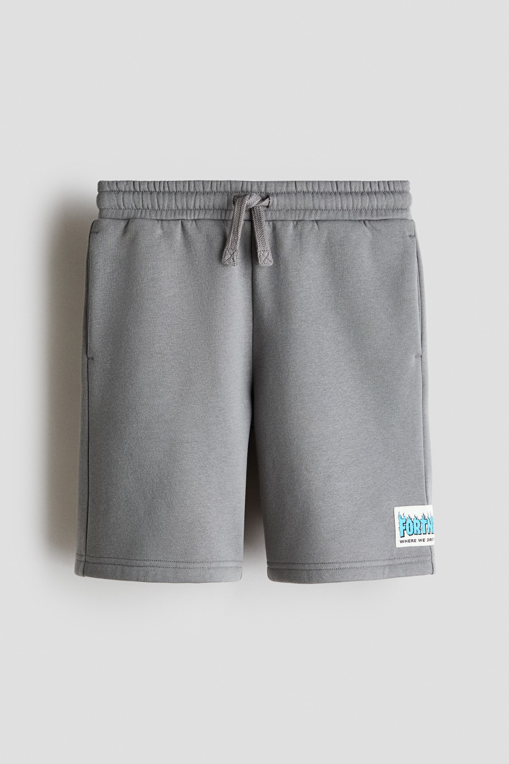 Visualizza l'immagine ingrandita: I pantaloncini grigi sono presentati da una vista frontale, con una cintura elastica dotata di un cordoncino grigio annodato. Sono visibili le tasche laterali e una toppa rettangolare con la scritta "Fortnite" in grafica blu e bianca con il testo "where we dro" è cucita sulla parte inferiore della gamba destra.