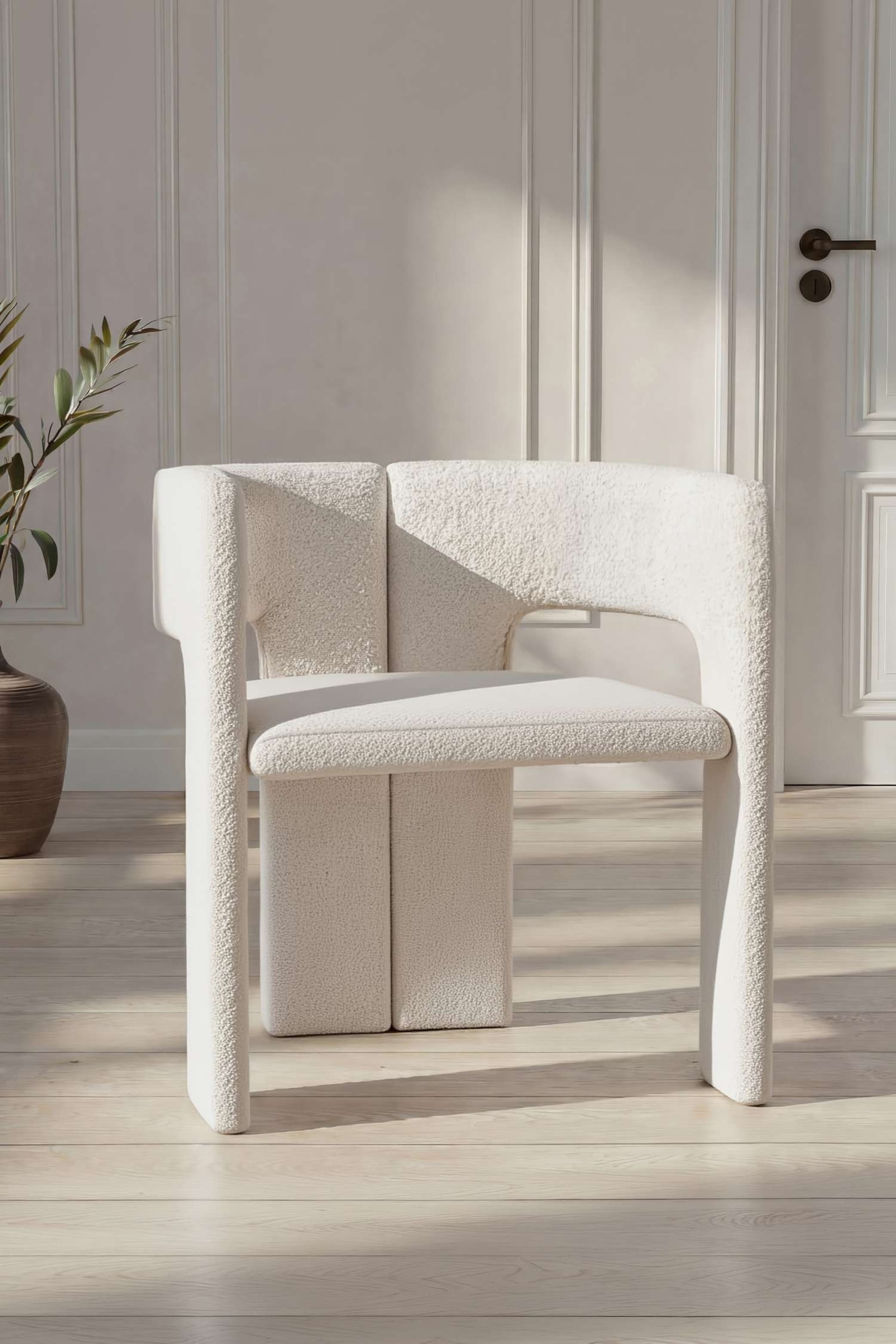 Ver imagen más grande: La silla de comedor Kristin presenta un diseño escultórico y moderno, tapizado en una tela bouclé texturizada de color crema, mostrada desde un ángulo de tres cuartos. Destaca por un respaldo curvado que envuelve el cuerpo con reposabrazos integrados y un asiento acolchado sostenido por patas gruesas de tipo columnar.