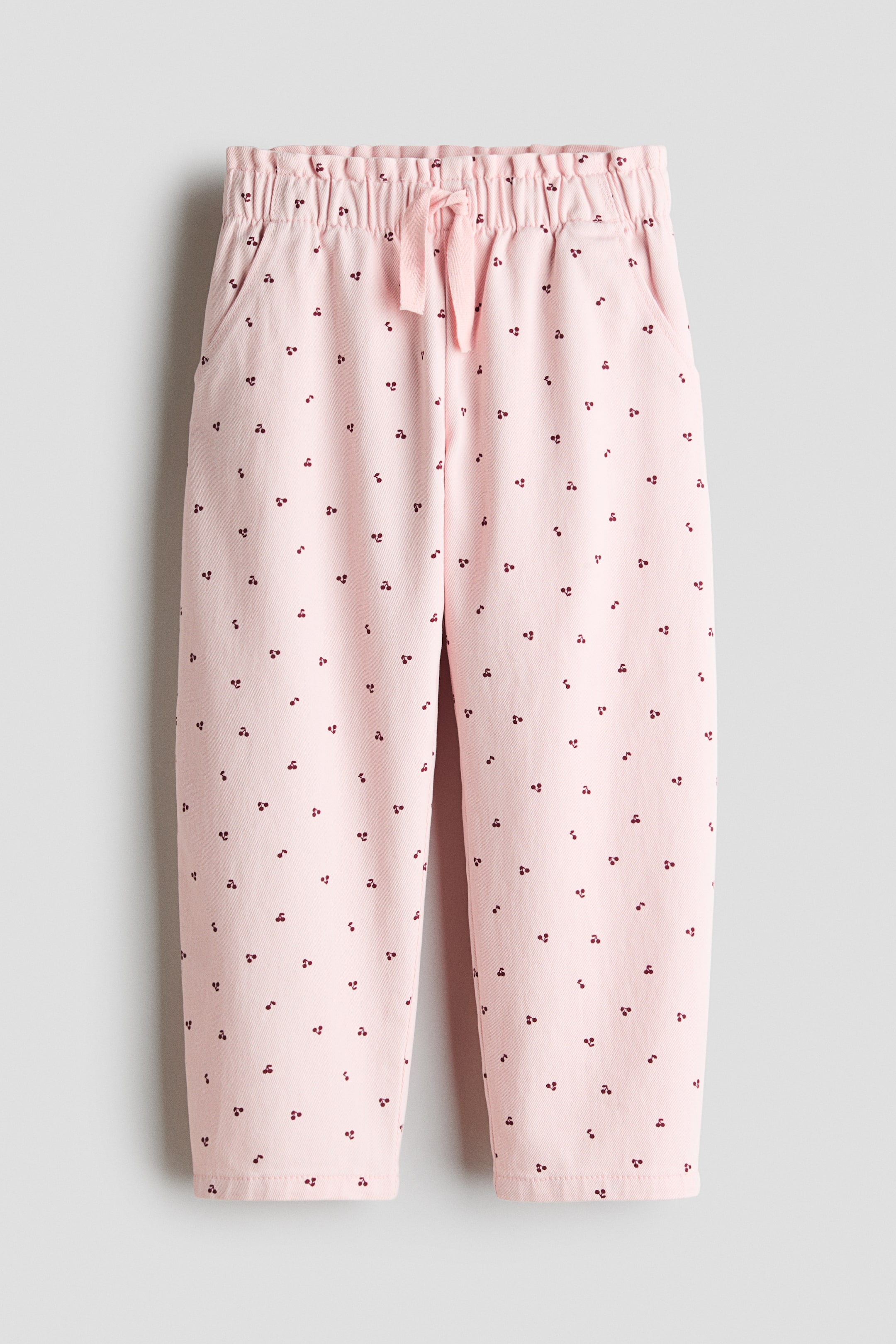 View larger image: Ang mga pink na pantalon ay may elastikong baywang na may pink na tali sa harap. Ang mga pantalon ay may buong disenyo ng maliliit na dark red na cherry motifs at dalawang bulsa sa gilid.