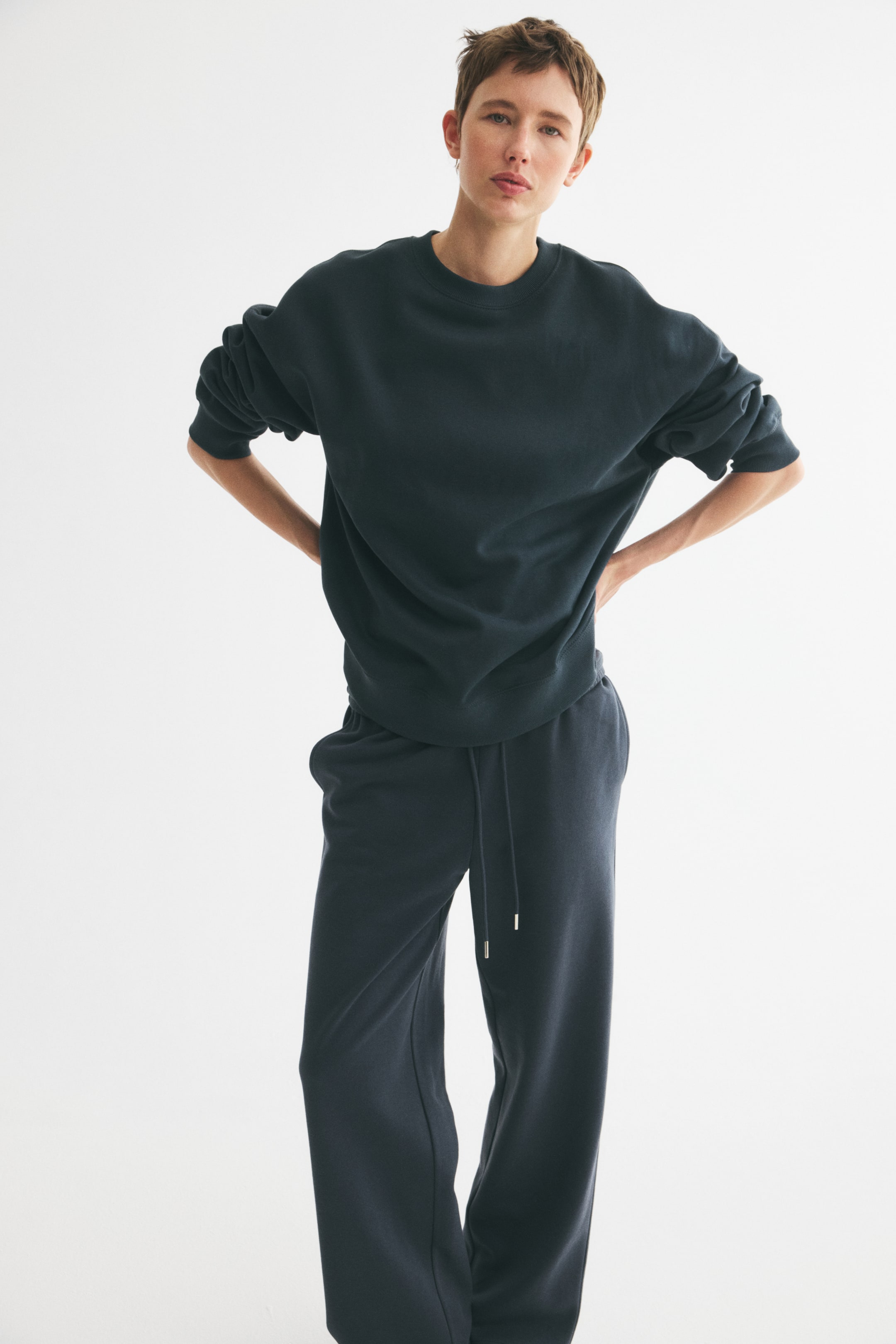View larger image: The model is wearing an oversized top crafted from dark sweatshirt fabric. It features a round neckline with ribbed trim, dropped shoulders, and long sleeves with ribbing at both the cuffs and hem. The model stands with hands on hips, showcasing the top paired with matching dark sweatpants.