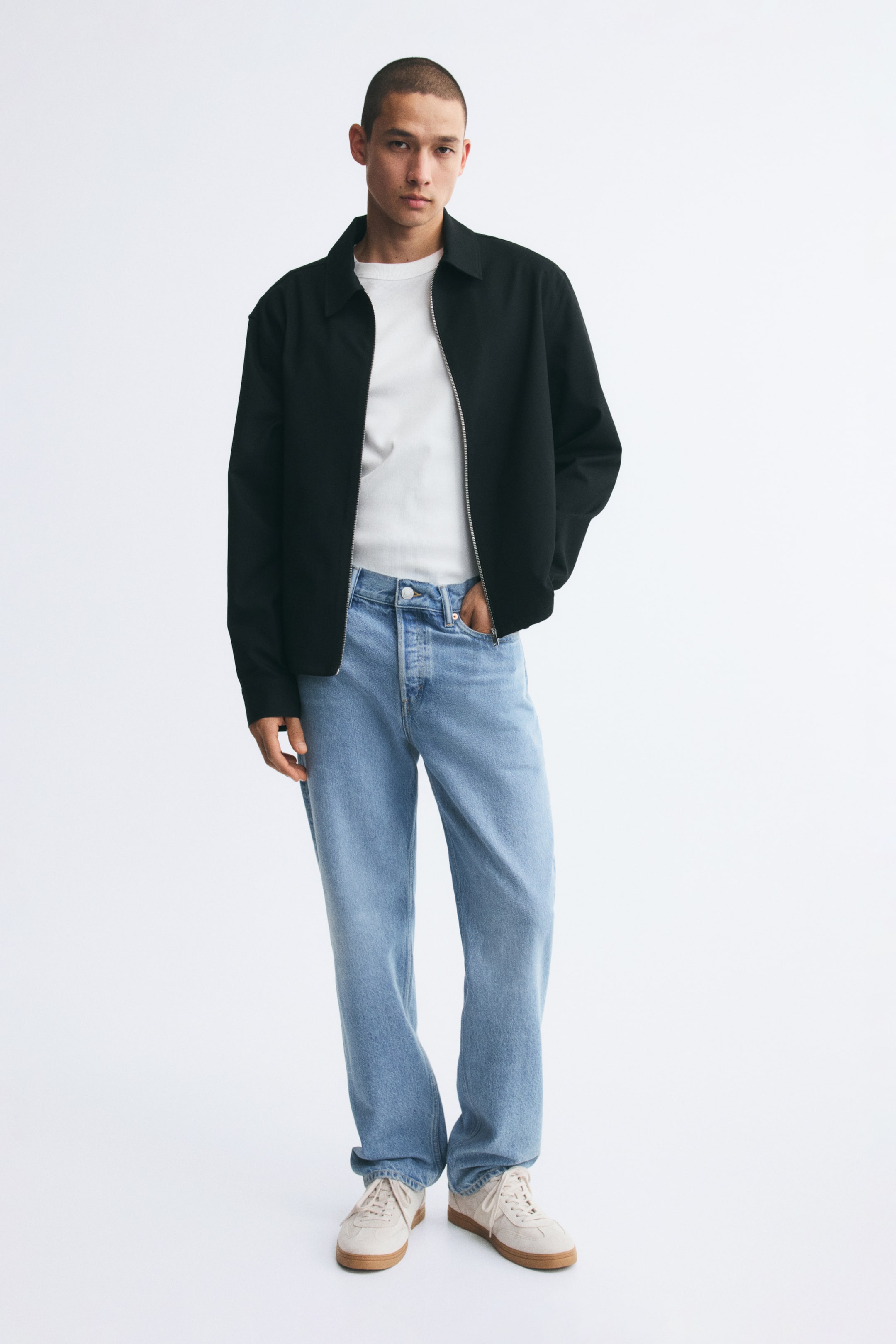 View larger image: The model is wearing straight-leg jeans in a light blue wash, with one hand placed in a front pocket. The jeans are styled with a tucked-in white T-shirt, an open black jacket, and off-white trainers.