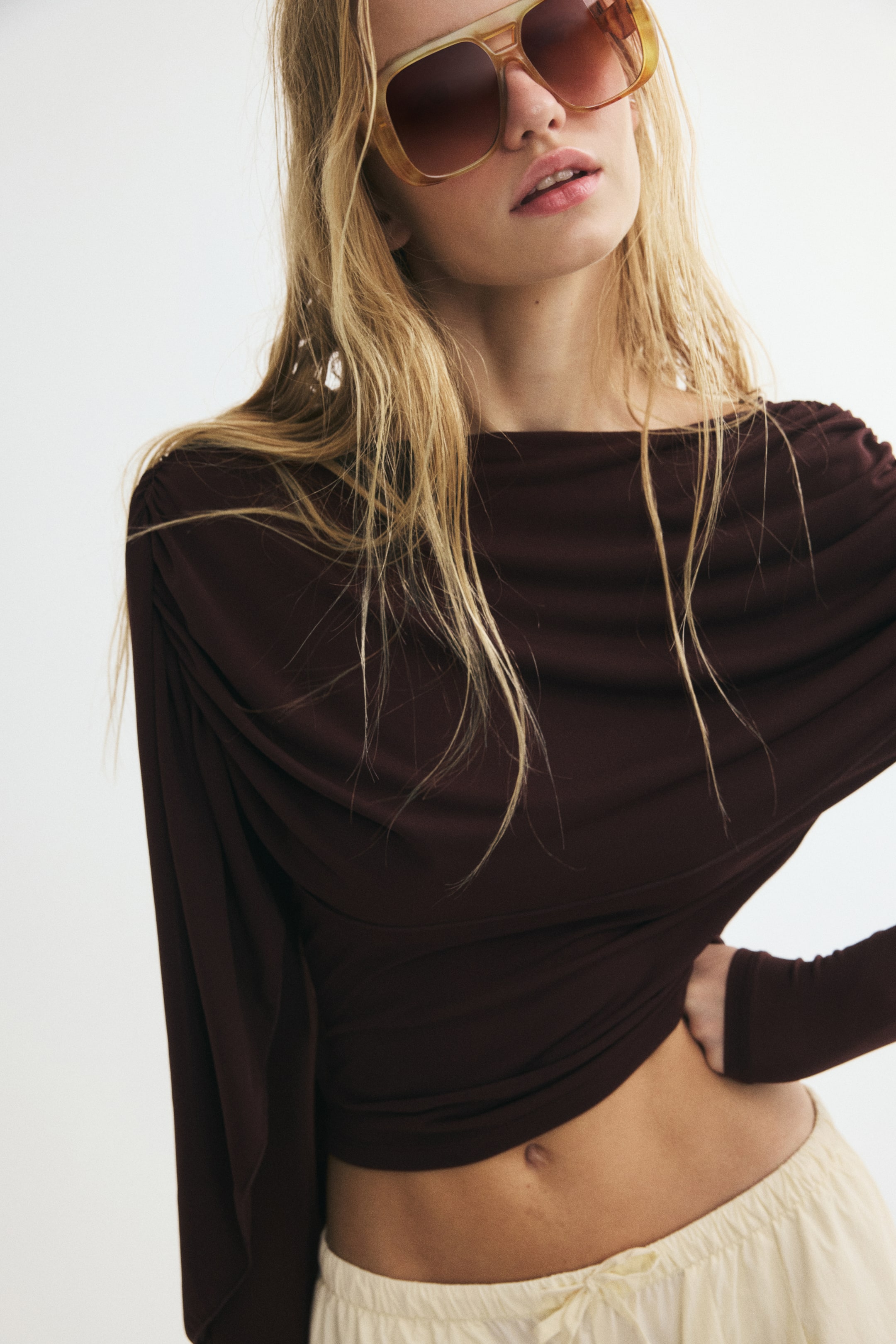 Xem ảnh lớn hơn: Người mẫu đang mặc một chiếc áo kiểu cape xếp drape màu nâu sẫm, với cổ rộng và phần vải mềm mại bất đối xứng buông nhẹ qua ngực. Chiếc áo tay dài được phối cùng quần màu kem và kính râm bản lớn, trong khi người mẫu tạo dáng với một tay đặt trên hông.