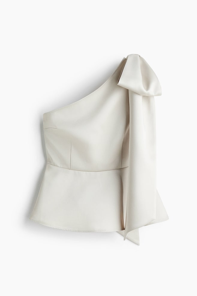 A light cream one-shoulder top in a smooth, lustrous fabric is visible from the front, featuring a large, structured bow on its right shoulder with a long, flowing sash. The top is presenting a fitted bodice with vertical darts and is flaring into a peplum hem. - 1