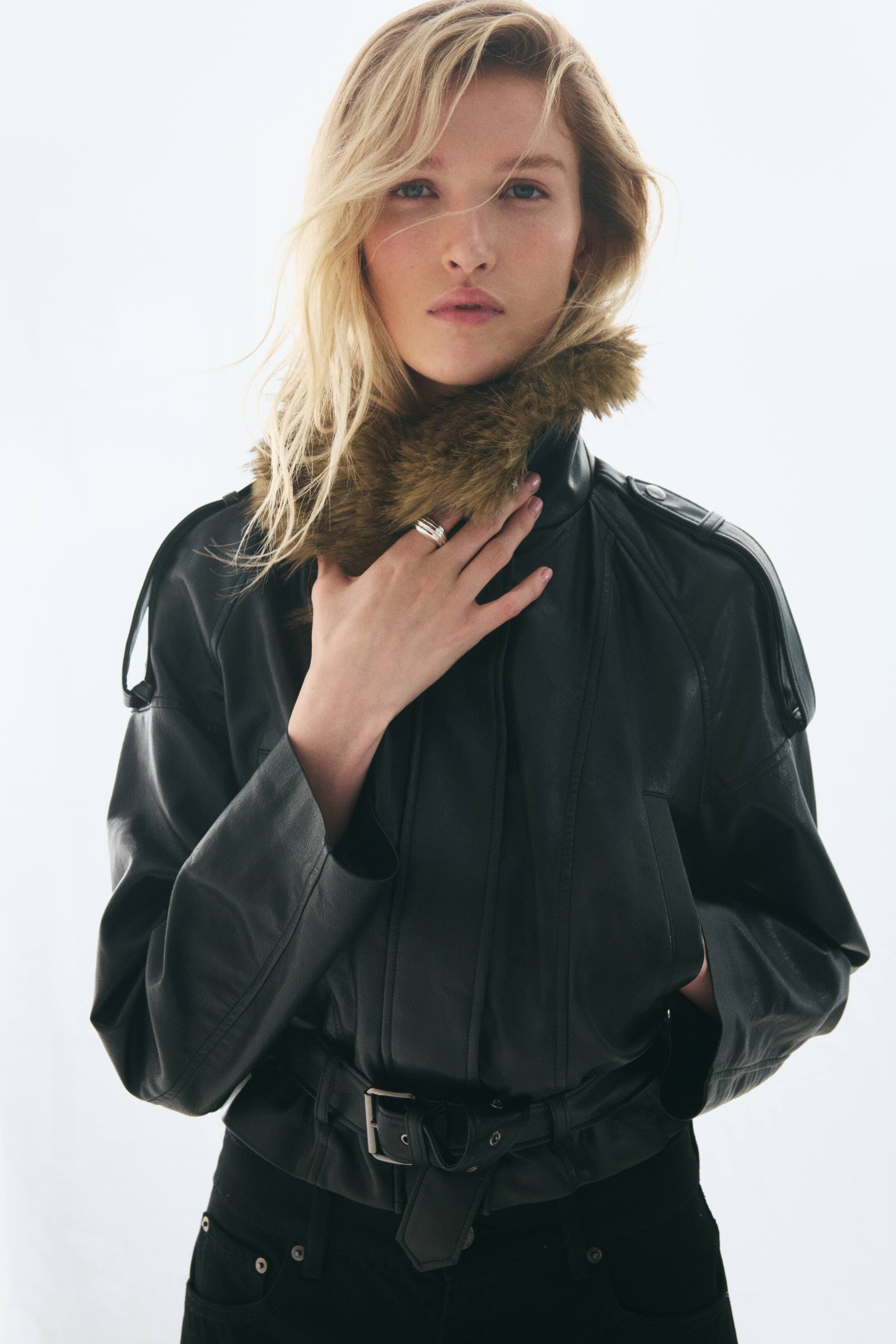 Größeres Bild ansehen: Vorderansicht einer kurzen schwarzen Jacke aus beschichtetem Stoff mit flauschigem braunem Kunstfellkragen und Schulterriegeln an überschnittenen Schultern. Die Jacke wird mit einem passenden, geschlossen getragenen Gürtel mit Metallschnalle am Saum kombiniert.