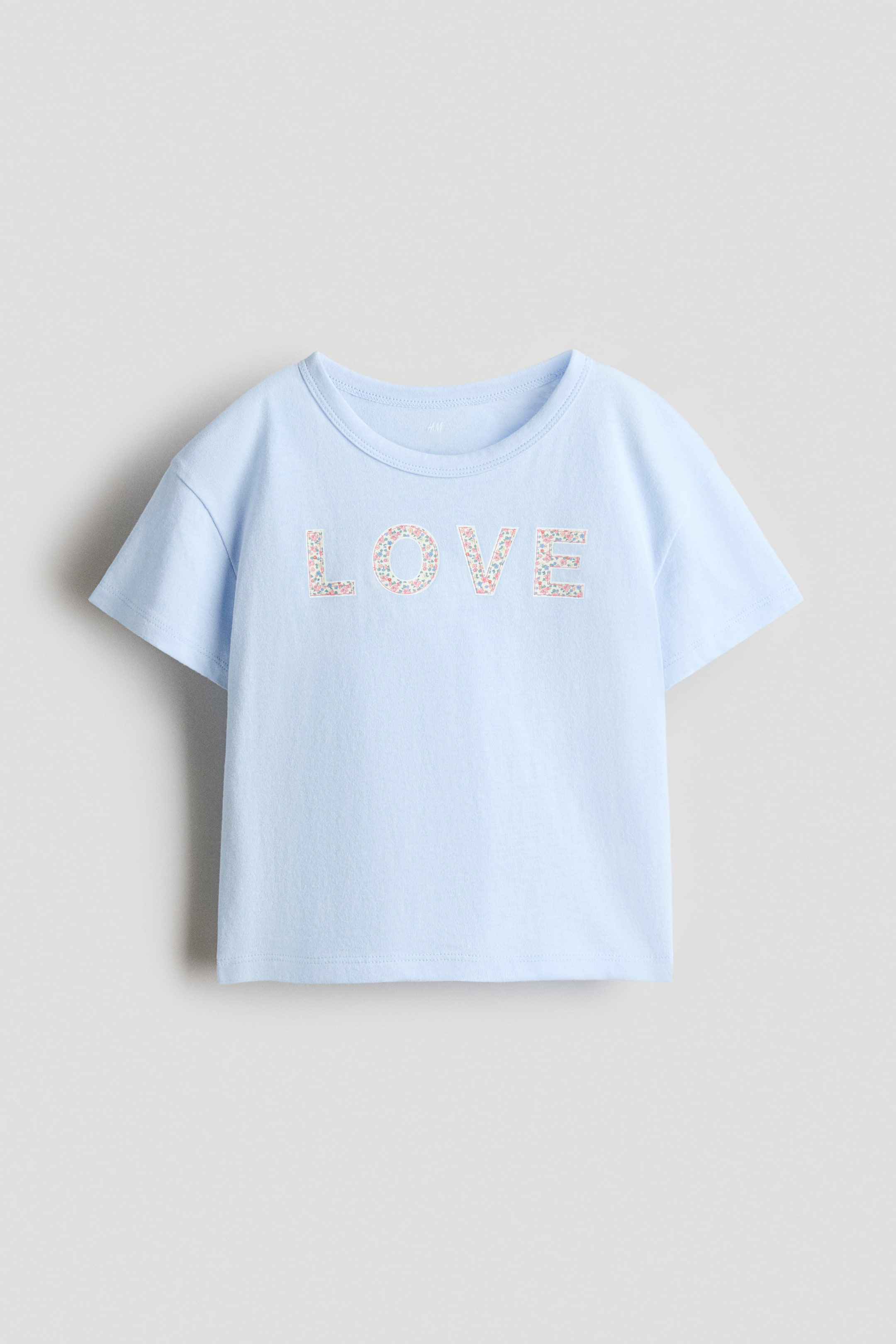View larger image: A light blue t-shirt is showcasing its front, with short sleeves and a round neckline. The word "LOVE" is displayed across the chest, adorned with a floral pattern featuring small pink, blue, and green elements.