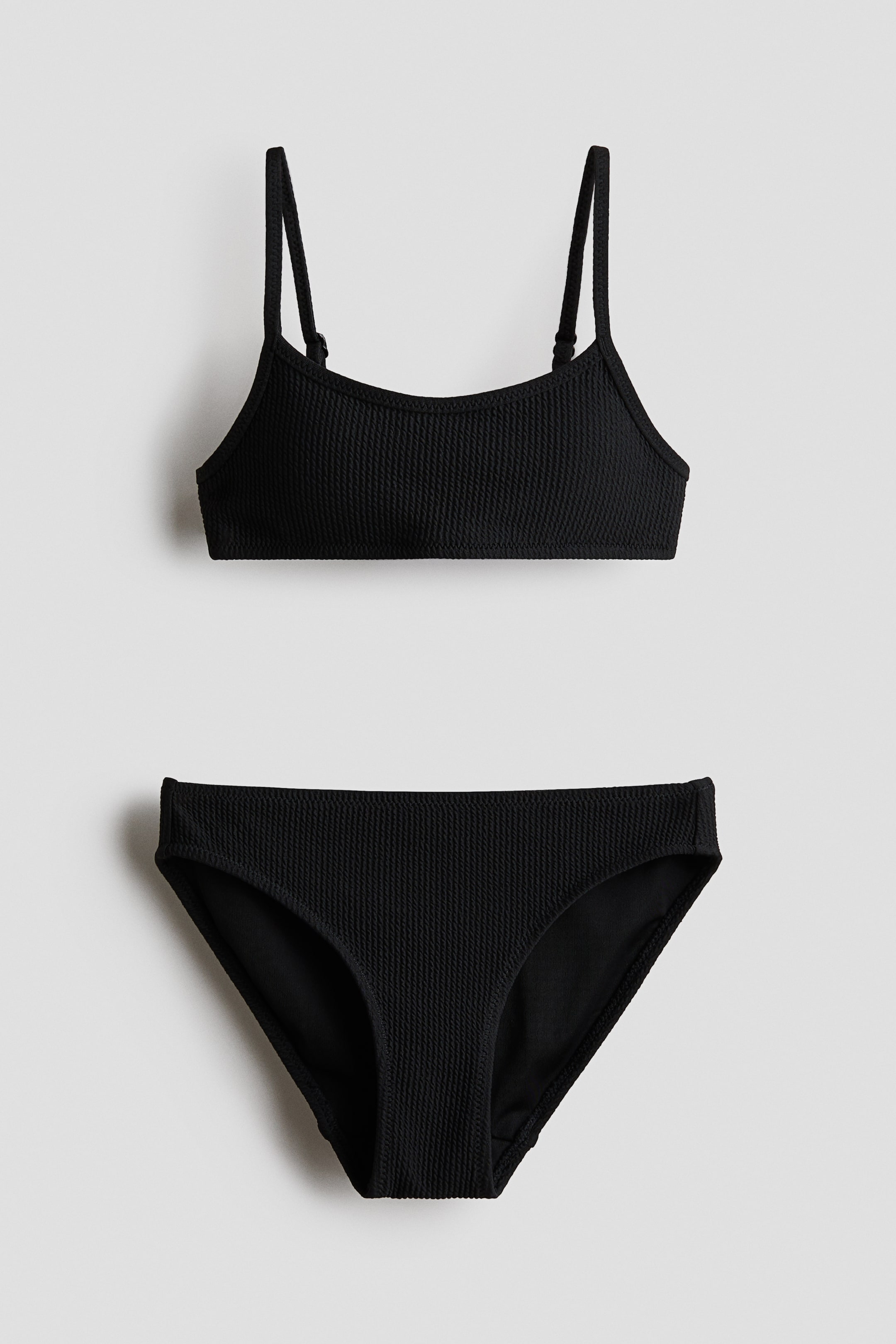 Agrandir l'image: Un maillot de bain deux pièces noir présente un tissu texturé, avec les deux pièces orientées face à la caméra. Le haut du bikini arbore un décolleté arrondi et des bretelles spaghetti ajustables, tandis que le bas de bikini assorti présente une coupe classique de slip.