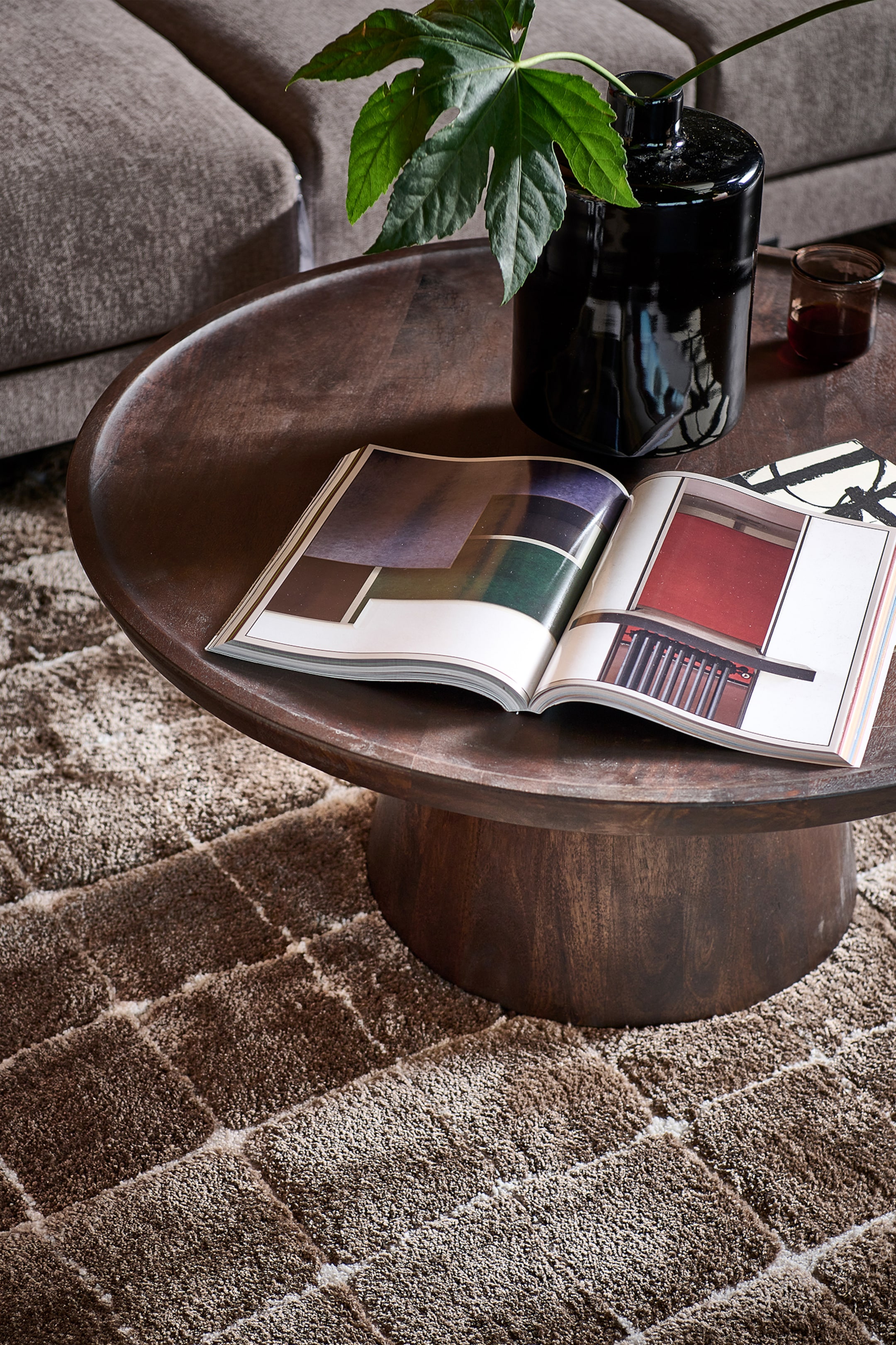 Agrandir l'image: Une table basse ronde en bois brun foncé présente un plateau en forme de plateau avec un rebord surélevé, vue d'un angle élevé. Elle repose sur une base cylindrique solide et expose un magazine ouvert sur sa surface.