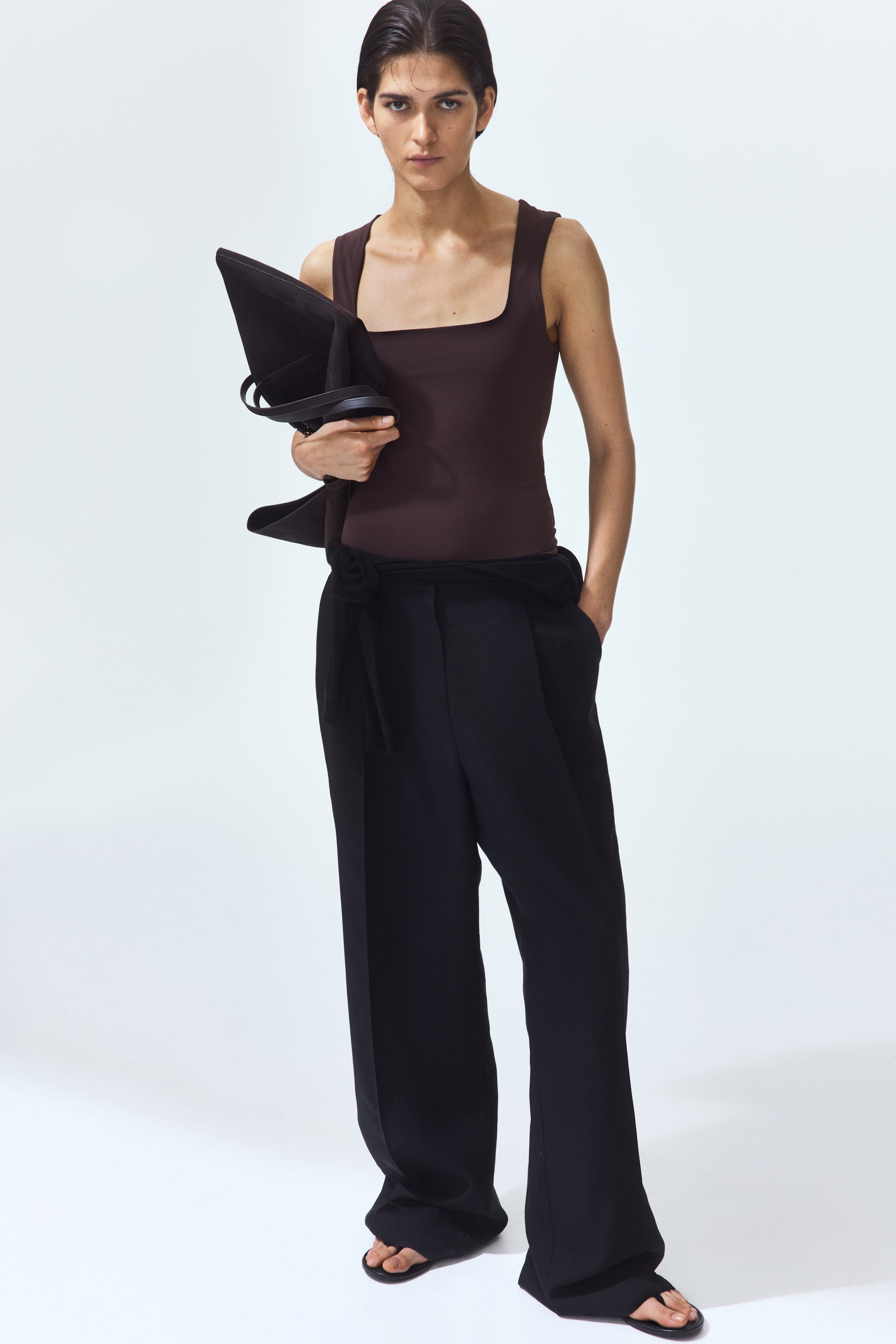 View larger image: The model is donning a dark brown fitted vest top crafted from heavy stretch jersey, featuring a square neckline and a straight-cut hem. This sleeveless top is paired with black trousers.
