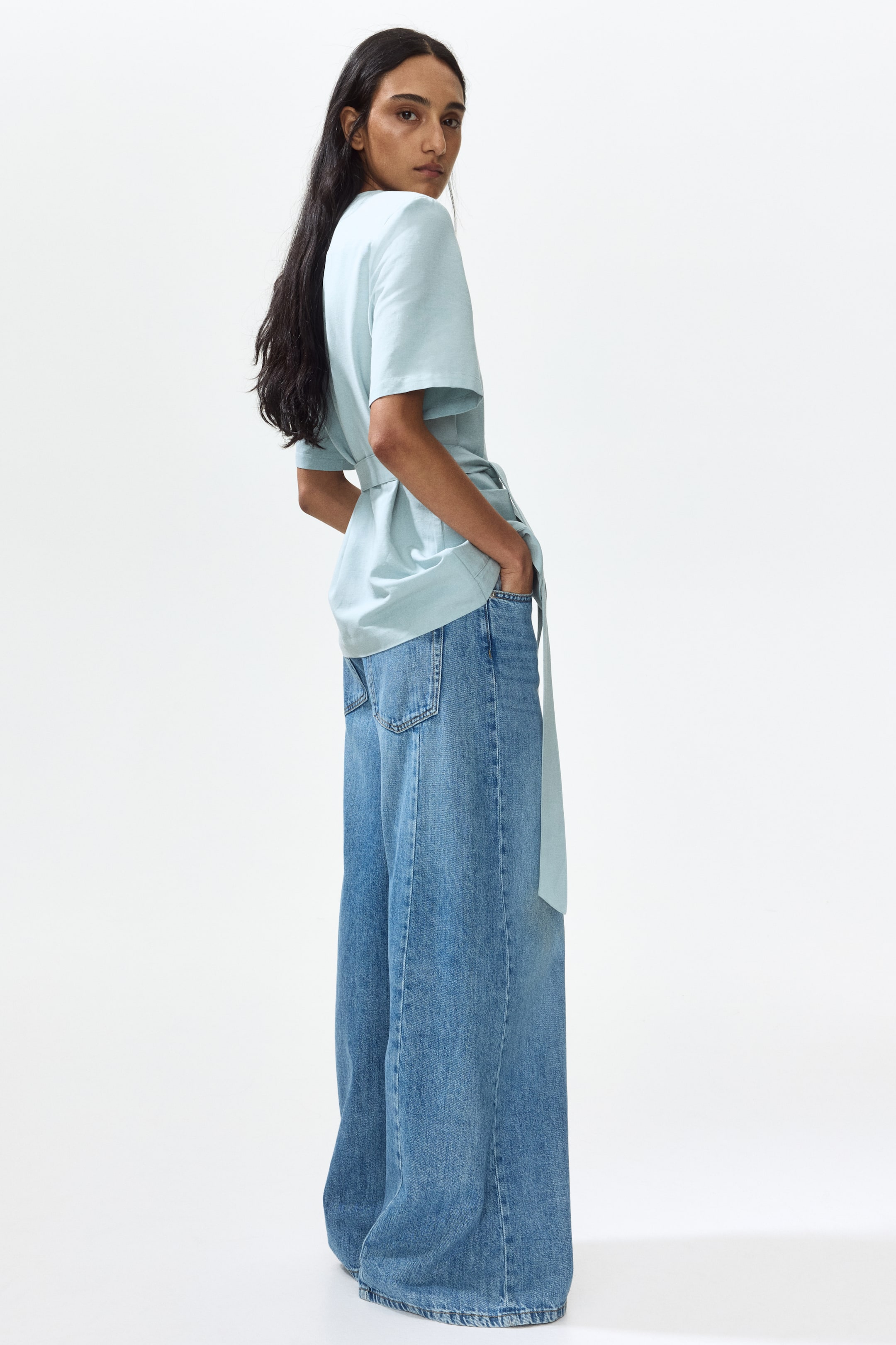 View larger image: A light blue short-sleeved top showcases a relaxed fit with a cinched tie-waist, its long tie elegantly draping over wide-leg blue jeans, as the model slightly turns to the left. The jeans feature a medium wash, a broad silhouette, and visible back pockets.