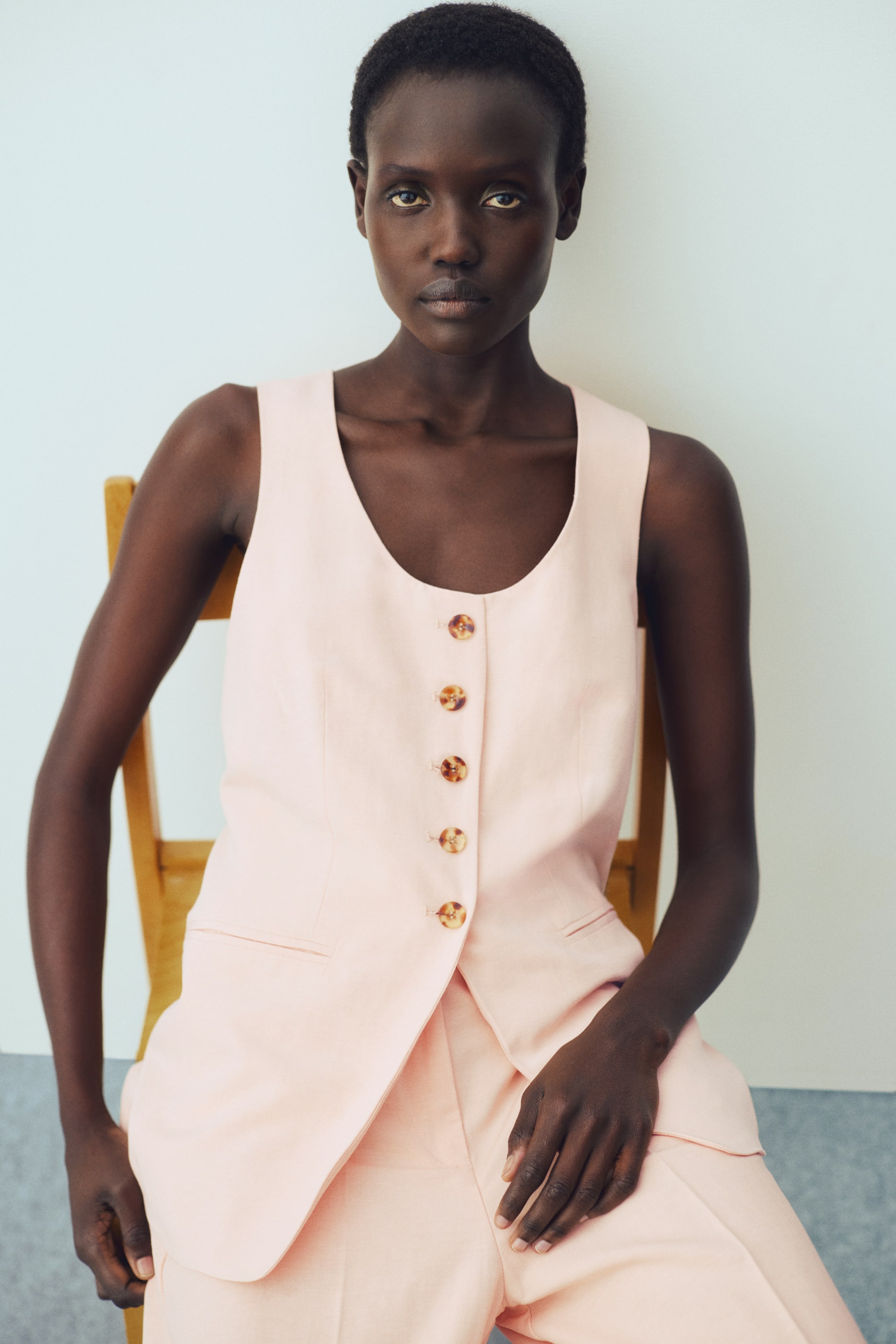 View larger image: A long, tailored waistcoat in woven fabric made from a linen blend is worn by the model. It features a scoop neckline, brown buttons down the front to the waist, a tapered waist with shaping darts that create a gently flared hem, and jetted front pockets. The model is seated, wearing the waistcoat with matching trousers.