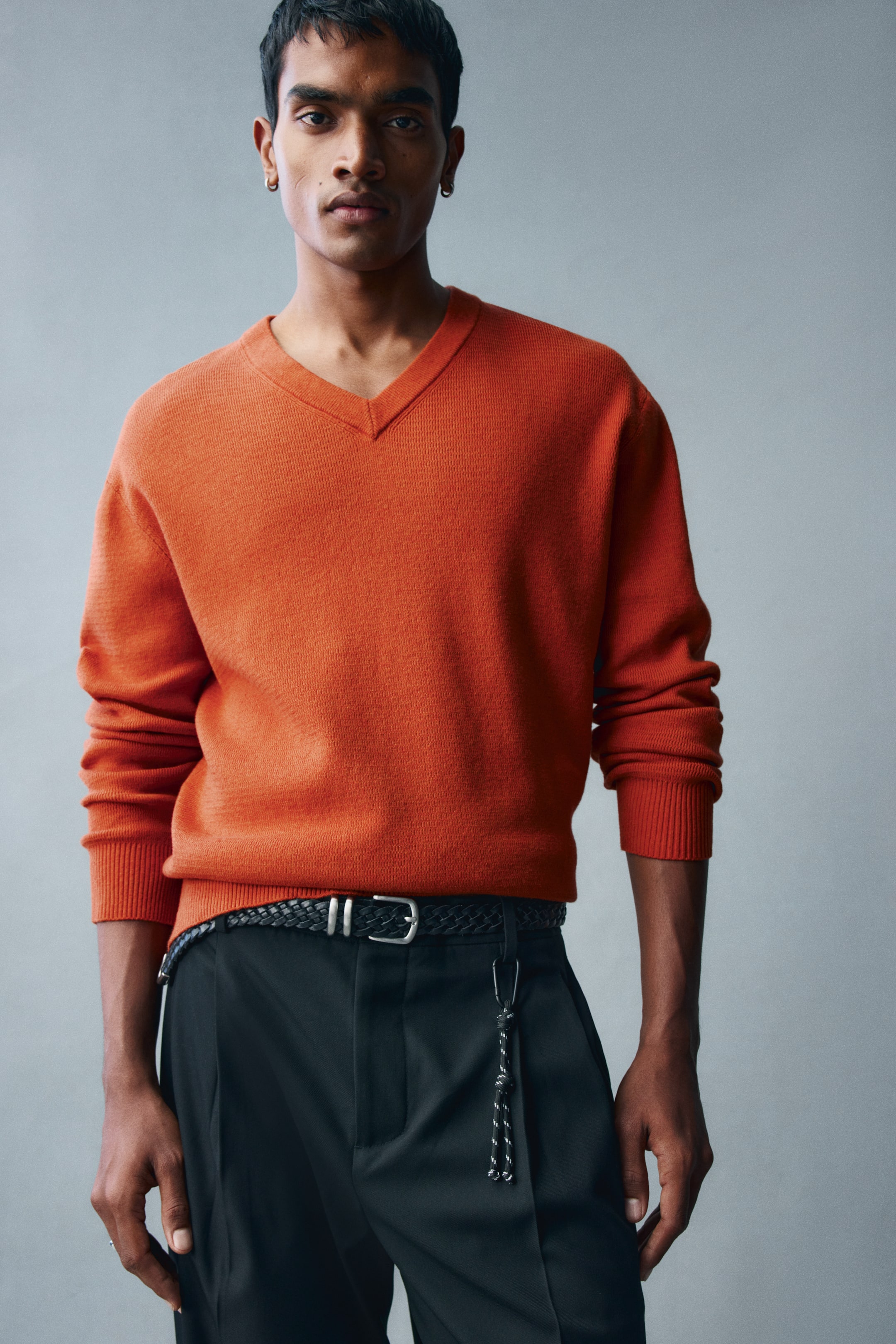 View larger image: The model is wearing an orange knit sweater featuring a relaxed fit, a ribbed V-neck, and ribbed trims at the cuffs and hem. The sweater is styled tucked into black trousers secured with a braided belt.