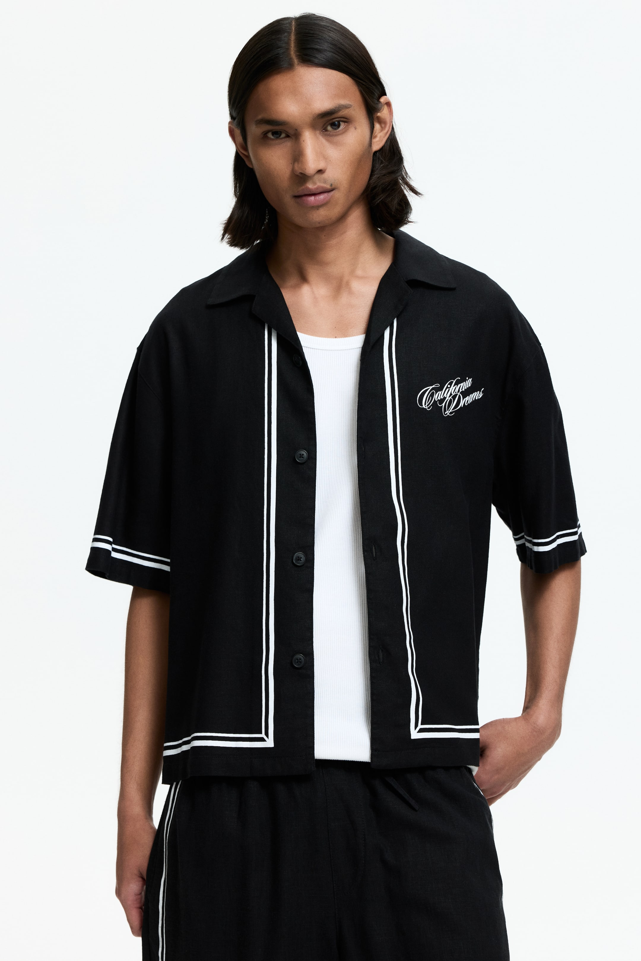View larger image: A black short-sleeve shirt, angled slightly to the right, showcases a camp collar with white piping outlining the open front, bottom hem, and sleeves. The left chest features white embroidered text reading 'California Dreams.' The shirt is styled open over a white ribbed tank top.