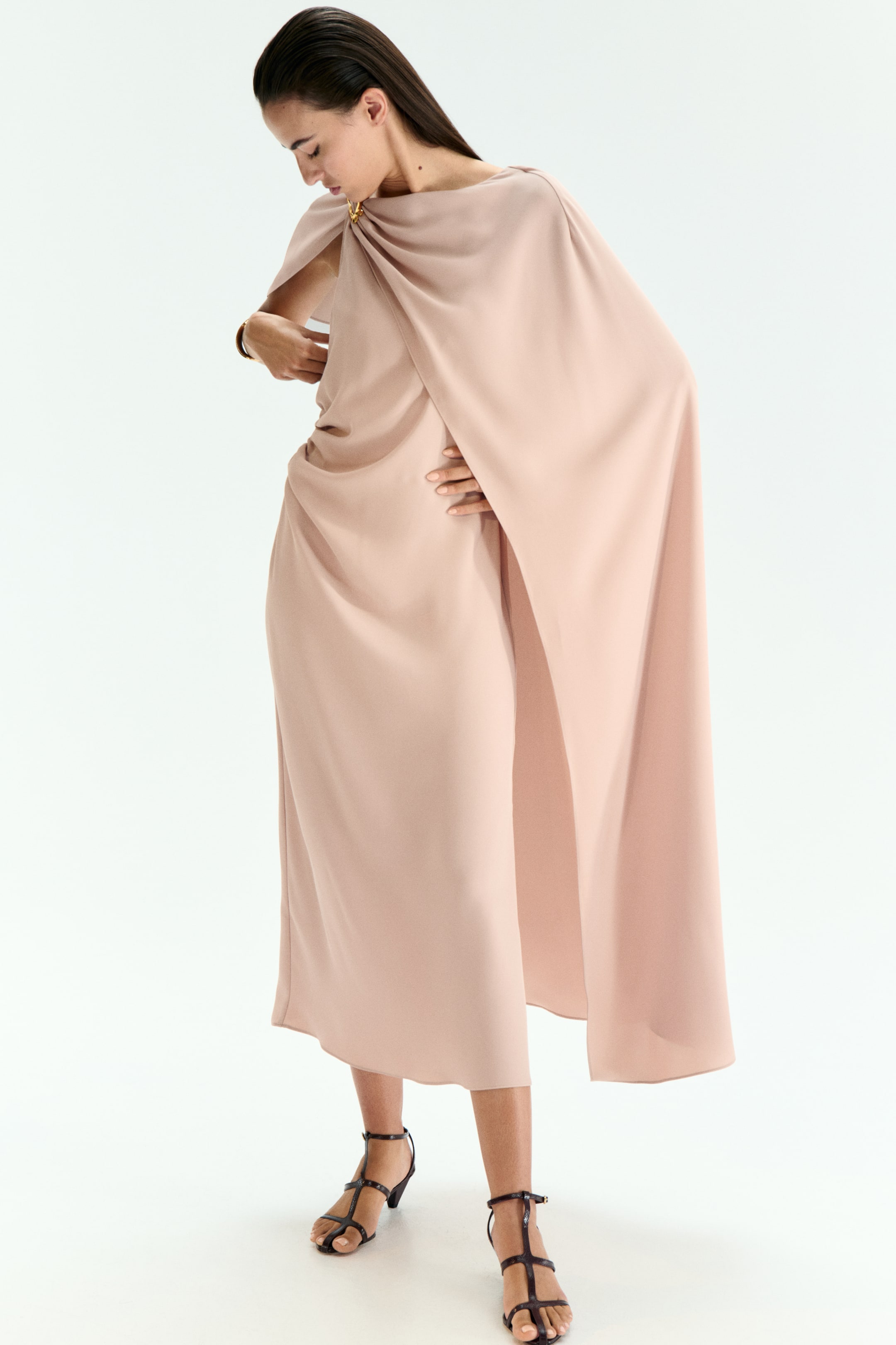 View larger image: A blush midi dress is featuring a draped silhouette, gathered at the right shoulder with a gold-toned circular clasp. A flowing cape is extending from the clasp, while soft fabric folds are draping around the waist and hips, falling to a midi length.