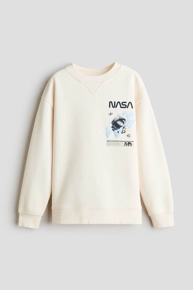 Una sudadera de cuello redondo en color blanco roto, vista de frente, presenta un cuello acanalado con inserción en V cosida, hombros caÃdos y puños y bajo acanalados. En el pecho izquierdo luce un logotipo negro de NASA sobre un gráfico de casco de astronauta superpuesto sobre letras estilizadas en azul claro. - 1