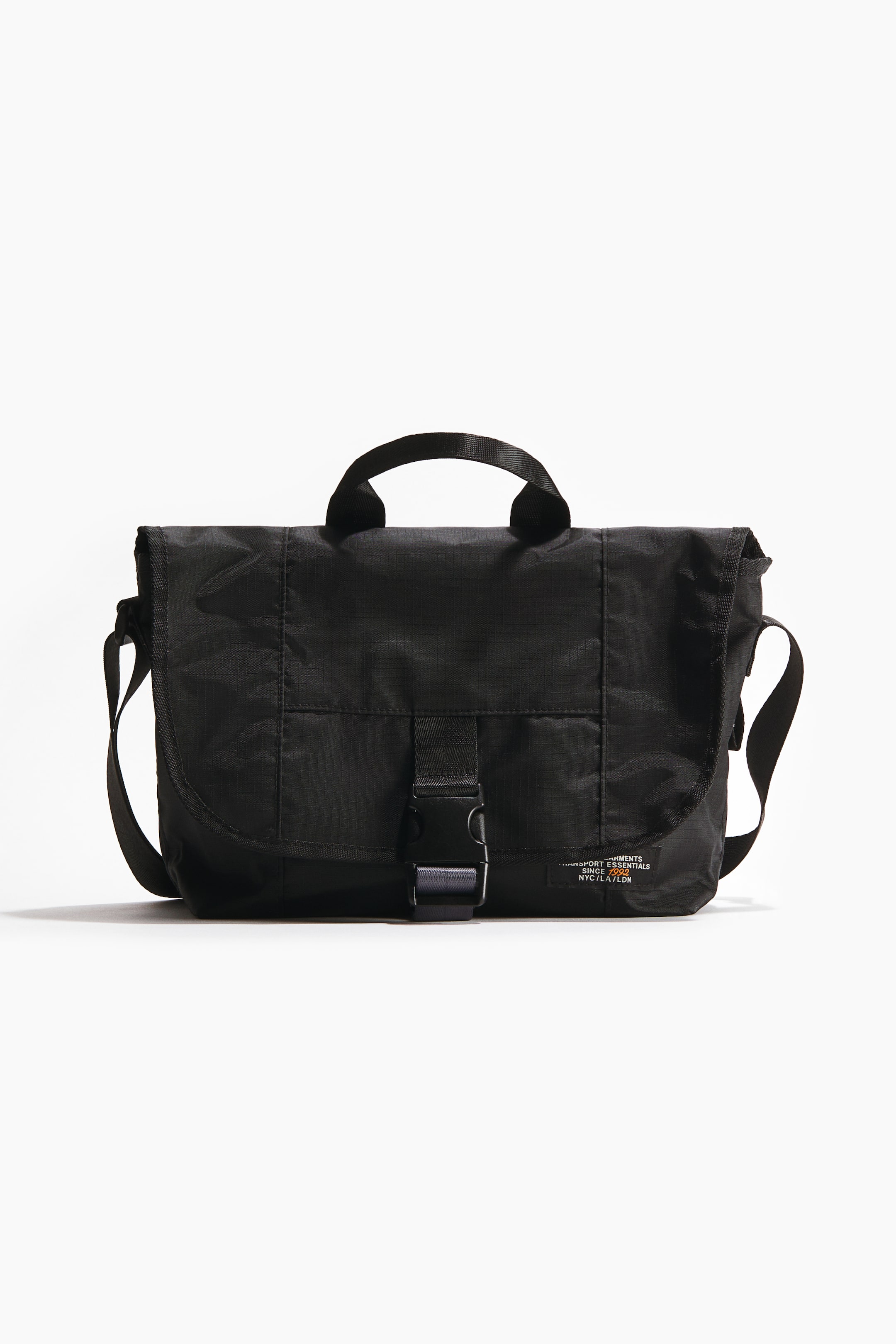 Ver imagen más grande: Un bolso bandolera negro se presenta de frente, con un cuerpo rectangular hecho de un tejido texturizado. Su solapa superior se cierra con una hebilla de plástico negro central y una correa de cinta, mientras que también son visibles un asa superior negra, una correa de hombro ajustable y una pequeña etiqueta de color claro con texto.