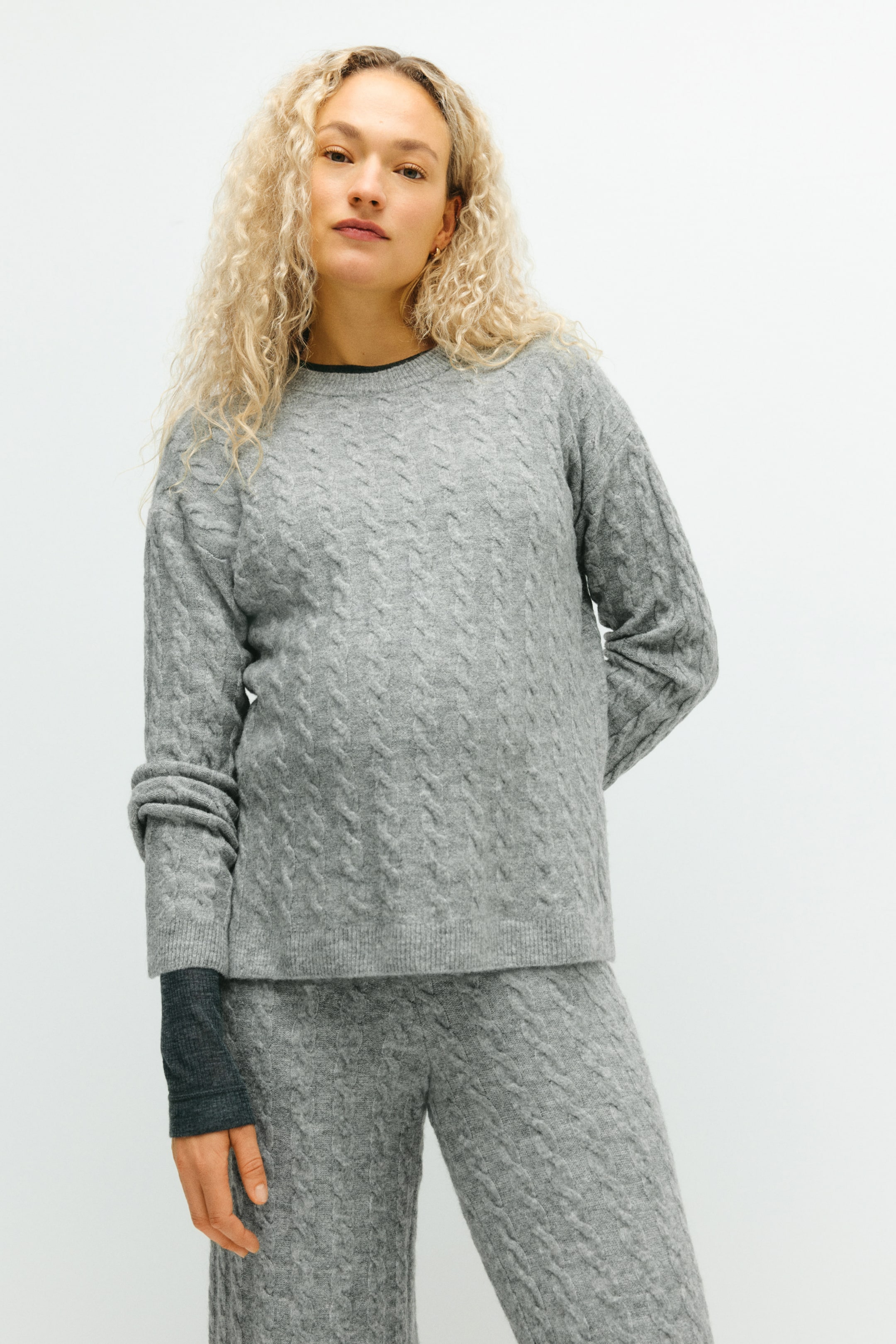 View larger image: A gray cable knit sweater and matching pants are being presented from the front. The relaxed-fit sweater is displaying a cable knit pattern throughout, featuring a crew neck, long sleeves with ribbed cuffs, and a ribbed hem, while the matching pants are also featuring the same cable knit pattern; a dark gray sleeve is visible layering under the sweater's left cuff.