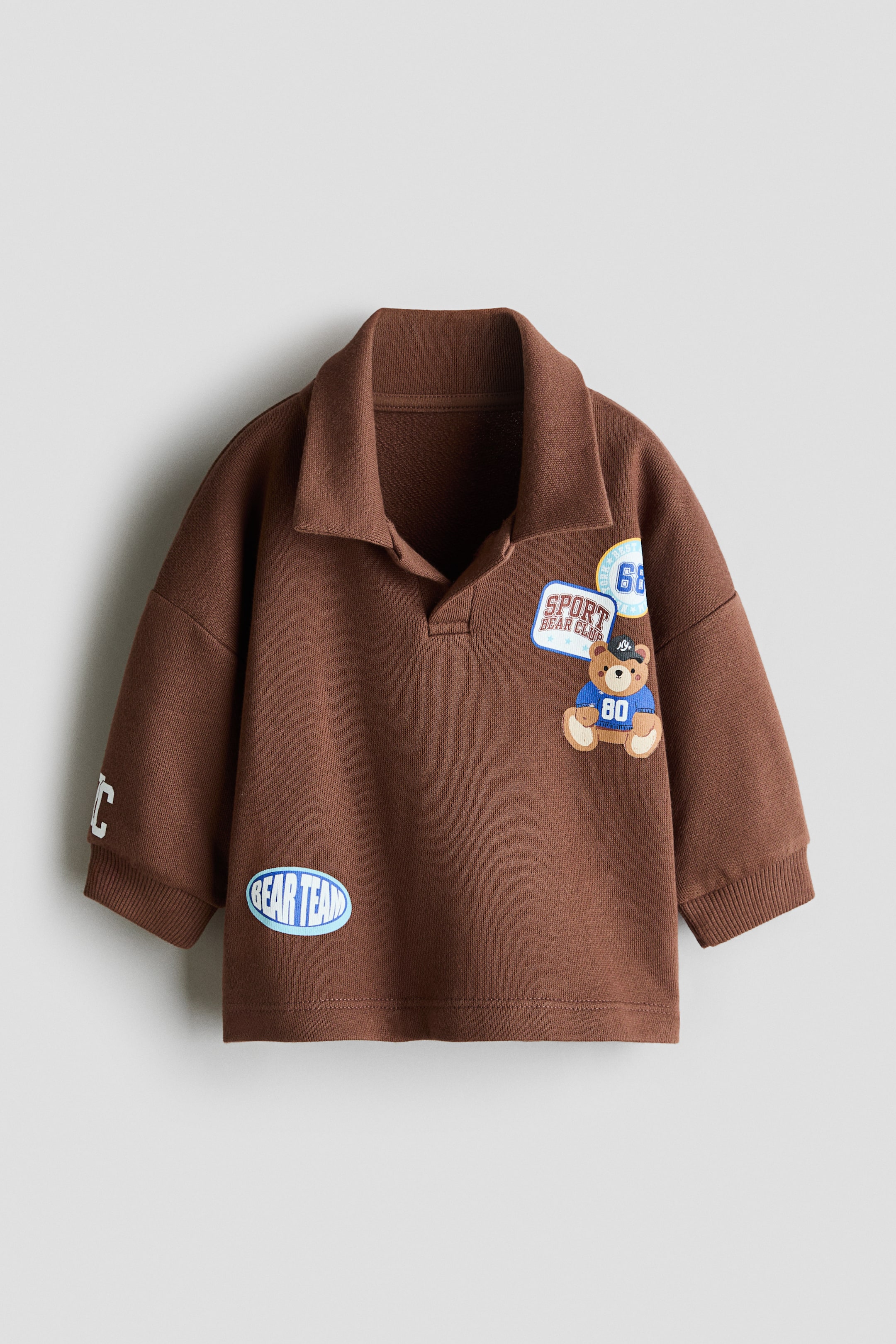 Visa större bild: En brun barnpolo-sweatshirt visas framifrån och framhäver en klassisk spread-krage, sänkta axlar och ribbade muddar. Den har flera varsity-liknande patchar, inklusive en tecknad nallebjörn på vänster bröst och en blå oval patch med texten "BEAR TEAM" nära nederkanten.