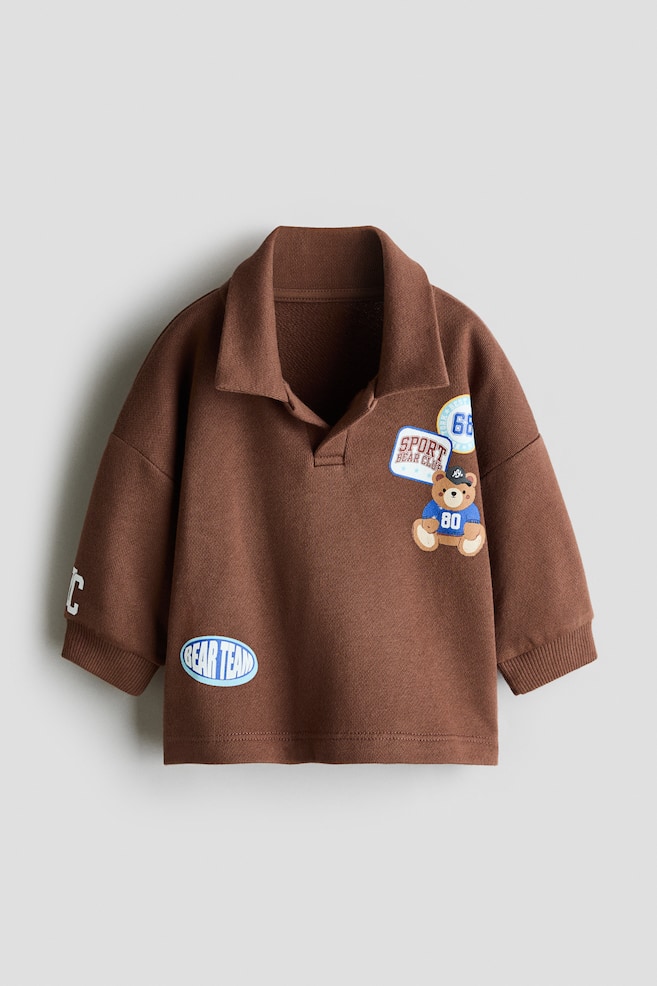 A brown childrenâs polo sweatshirt is shown front-on, featuring a classic spread collar, dropped shoulders, and ribbed cuffs. It displays multiple varsity-style patches, including a cartoon teddy bear on the left chest and a blue âBEAR TEAMâ oval patch near the bottom hem. - 1