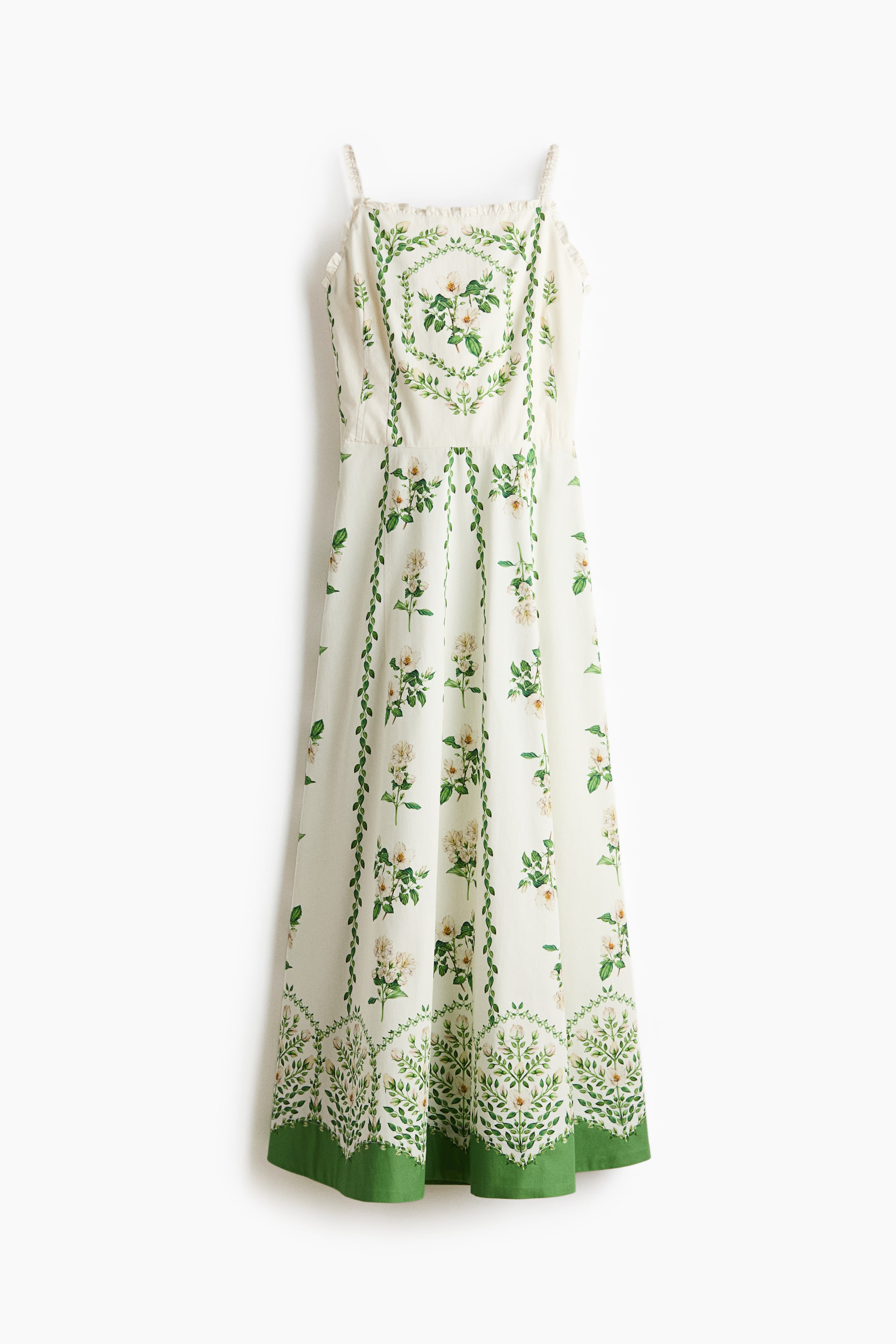 View larger image: A dress is hanging, facing front, showcasing an off-white fabric with a green and white floral and botanical print. It features thin spaghetti straps and a subtly ruffled square neckline. The dress body displays vertical leafy vine patterns with scattered white roses, and the hem presents a wide solid green border topped with a dense botanical frieze.