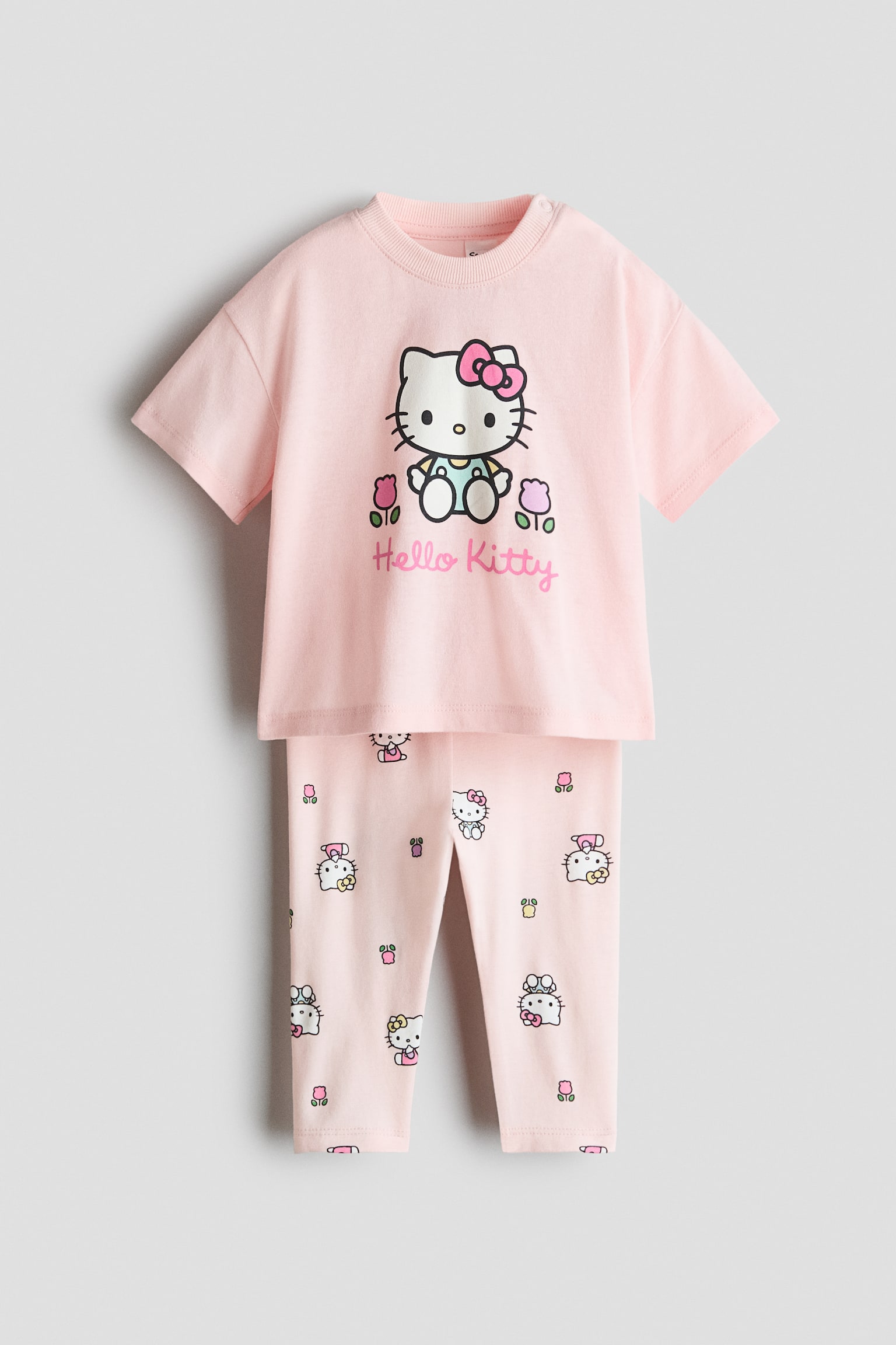 A light pink two-piece clothing set is shown from the front, featuring a short-sleeved t-shirt and matching pants. The t-shirt is displaying a large central Hello Kitty graphic with cursive text underneath, while the pants are featuring an all-over print of smaller Hello Kitty characters and flowers.