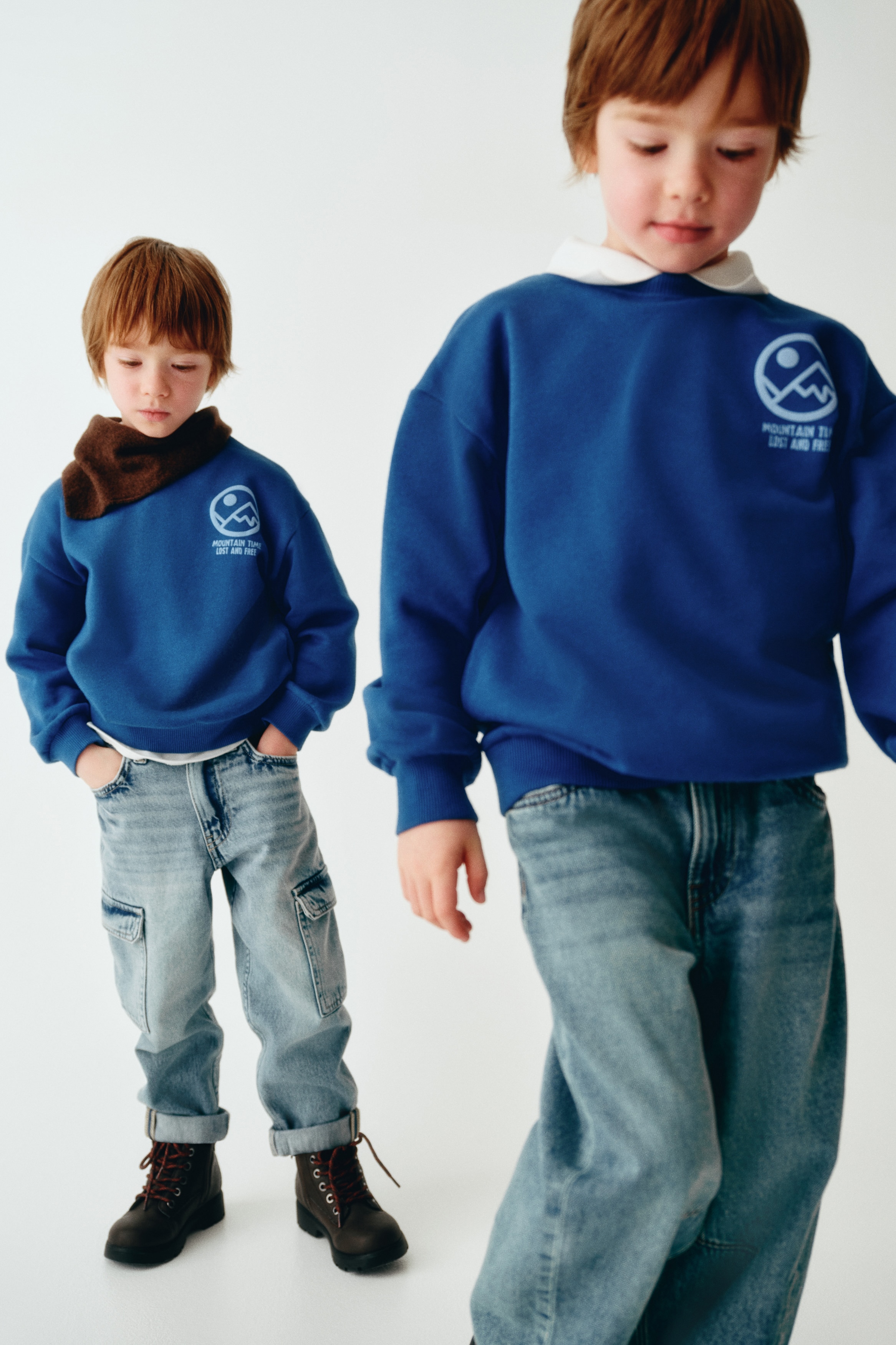 Egy kék sweatshirt elölnézetben látható, kerek, bordázott nyakkivágással, ejtett vállakkal és hosszú ujjakkal, bordázott mandzsettákkal. A sweatshirt bal mellrészén egy világoskék grafikai nyomat látható, amely egy kör alakú hegy motívumot és a „MOUNTAIN TIME LOST AND FREE” feliratot ábrázolja, a bordázott alsó szegély felett.