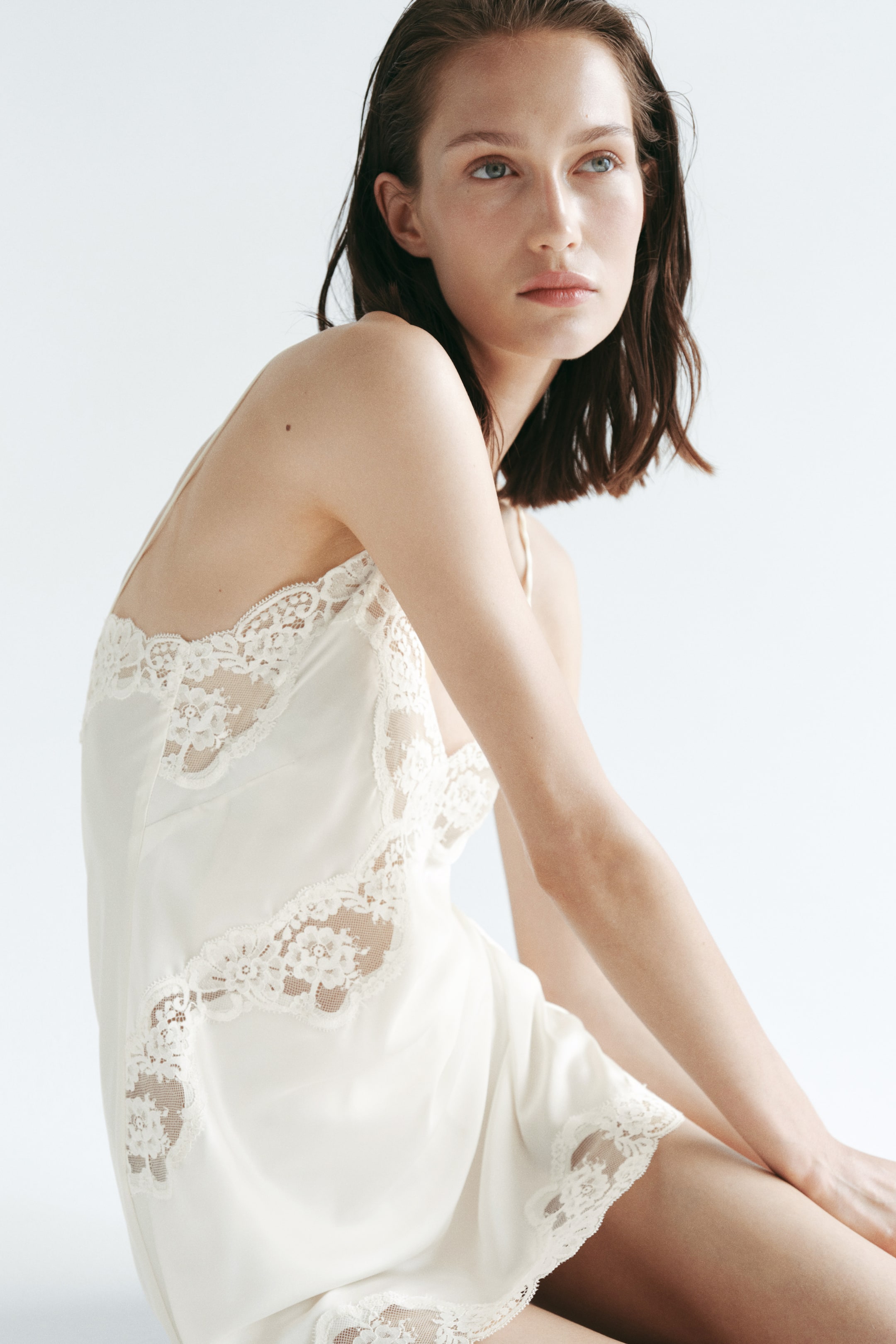 View larger image: The model is wearing an off-white, short, and fitted nightslip crafted from softly draping satin. It showcases a V-neckline, delicate spaghetti straps, and intricate floral lace trims and insets adorning the neckline and body.