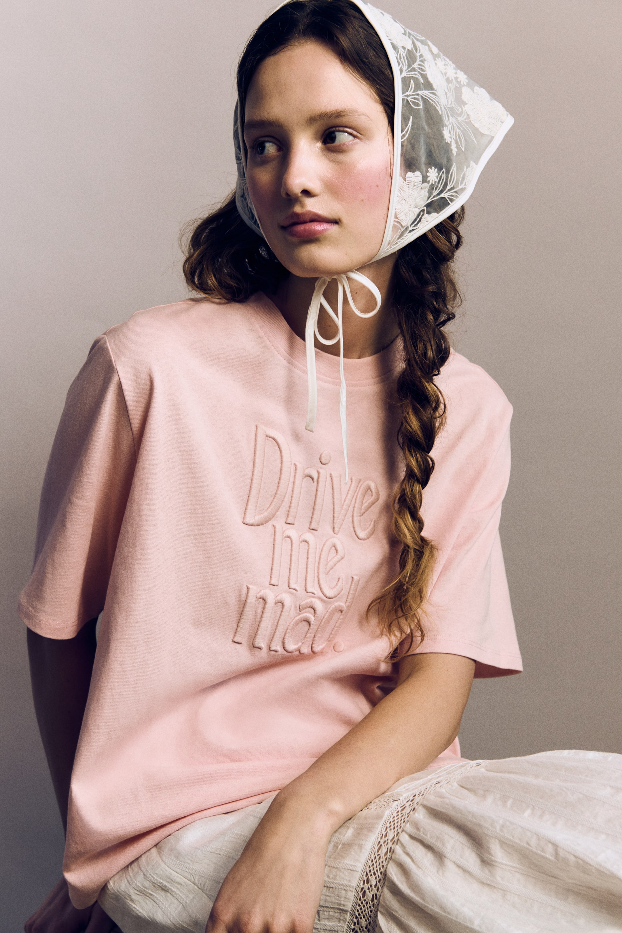 הצגת תמונה גדולה יותר: חולצת טי בגזרה רחבה בצבע ורוד בהיר נלבשת על ידי הדוגמנית, עם קו צוואר עגול עם גימור צלעות, כתפיים שמוטות ושרוולים קצרים. מוטיב רקום "Drive me mad" נראה בחזית, והחולצה מונחת מעל חצאית פליסה בצבע בהיר.