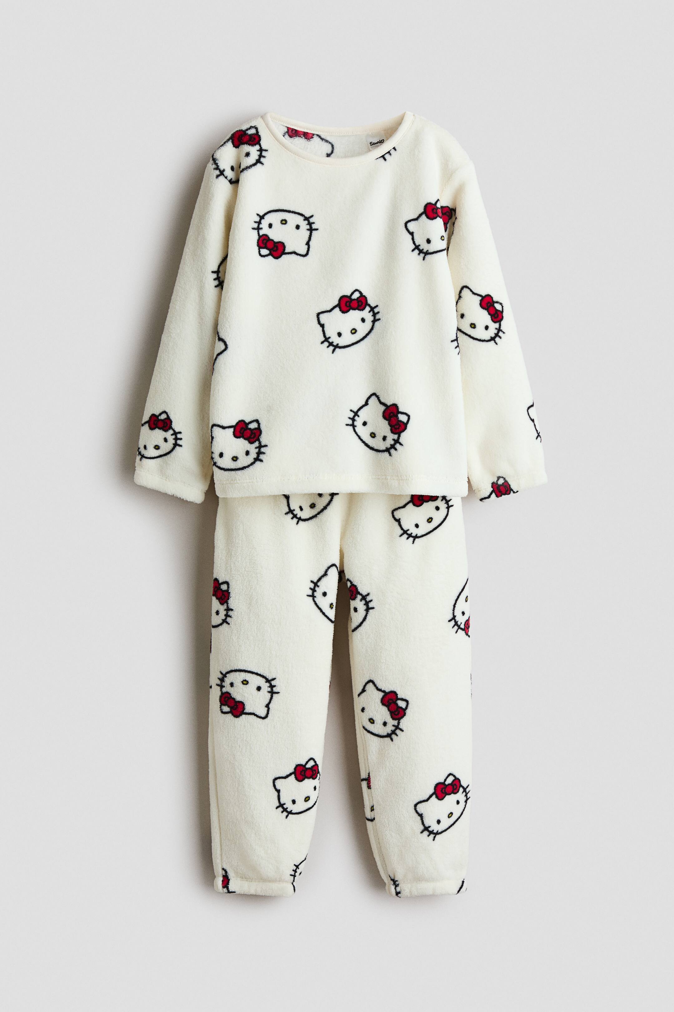 Visualizza immagine più grande: Un pigiama in pile color crema è presentato, caratterizzato da un motivo ripetuto di volti di Hello Kitty con contorno nero e fiocchi rossi. Il комплект comprende una maglia a maniche lunghe con girocollo e pantaloni lunghi coordinati, entrambi realizzati in un materiale morbido e soffice.