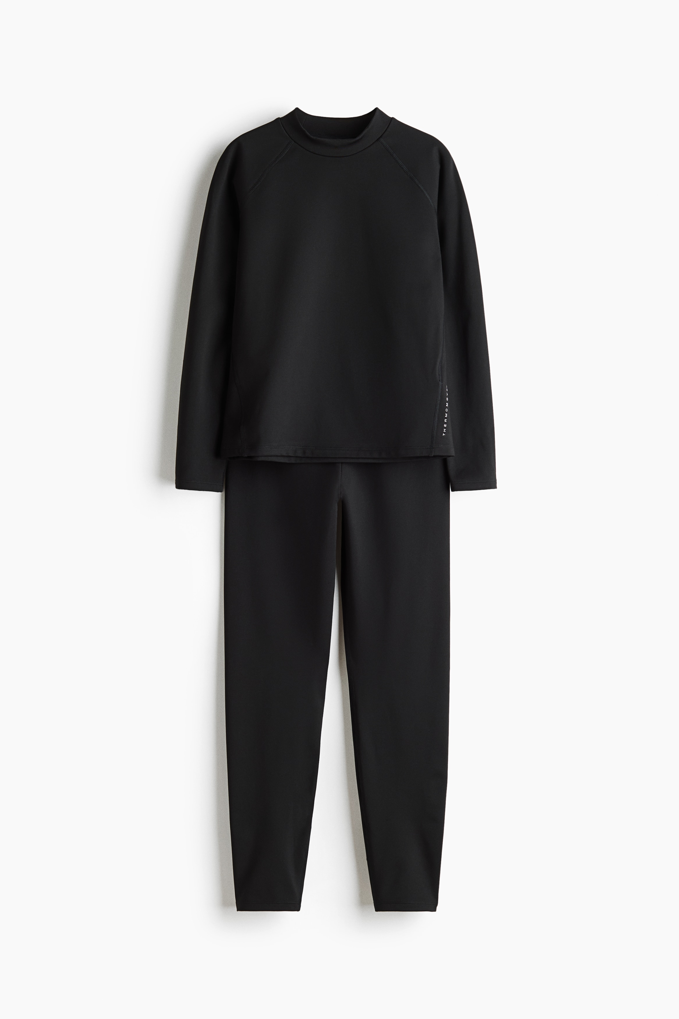 Un model stÄ Ã®n picioare, orientat spre faÈÄ, purtÃ¢nd un top negru cu mÃ¢neci lungi Èi guler tip mock-neck, pantaloni scurÈi negri asortaÈi, purtaÈi peste colanÈi negri, o cÄciulÄ neagrÄ cu un design curbat argintiu Èi pantofi sport albi cu detalii argintii. Ãn mÃ¢na dreaptÄ, acoperitÄ cu mÄnuÈÄ, Èine de Èireturi o pereche de ghete de fotbal negre.